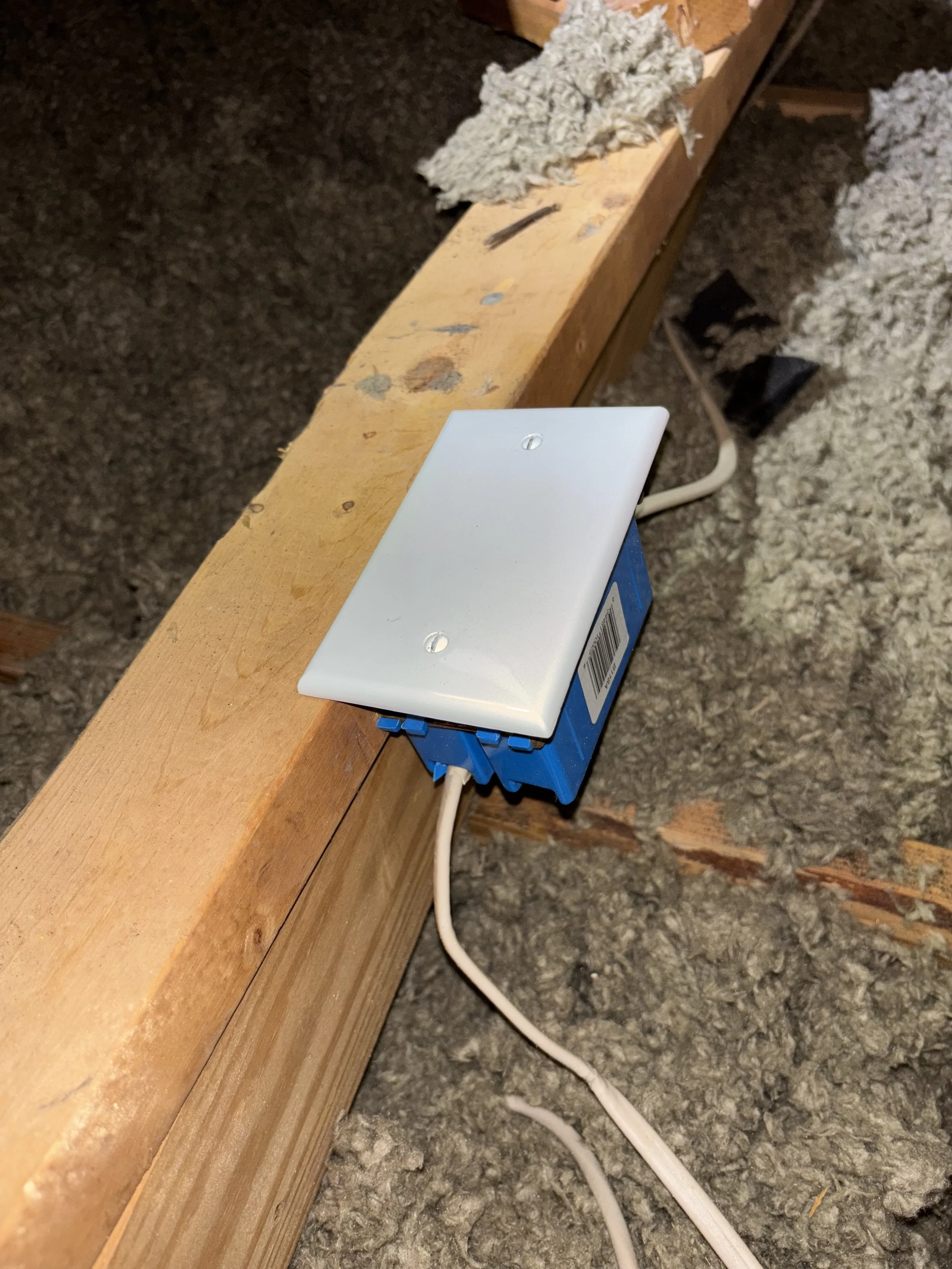 Electrical box with a white cover mounted on a wooden beam, with wires leading into it, in a construction or attic space.
