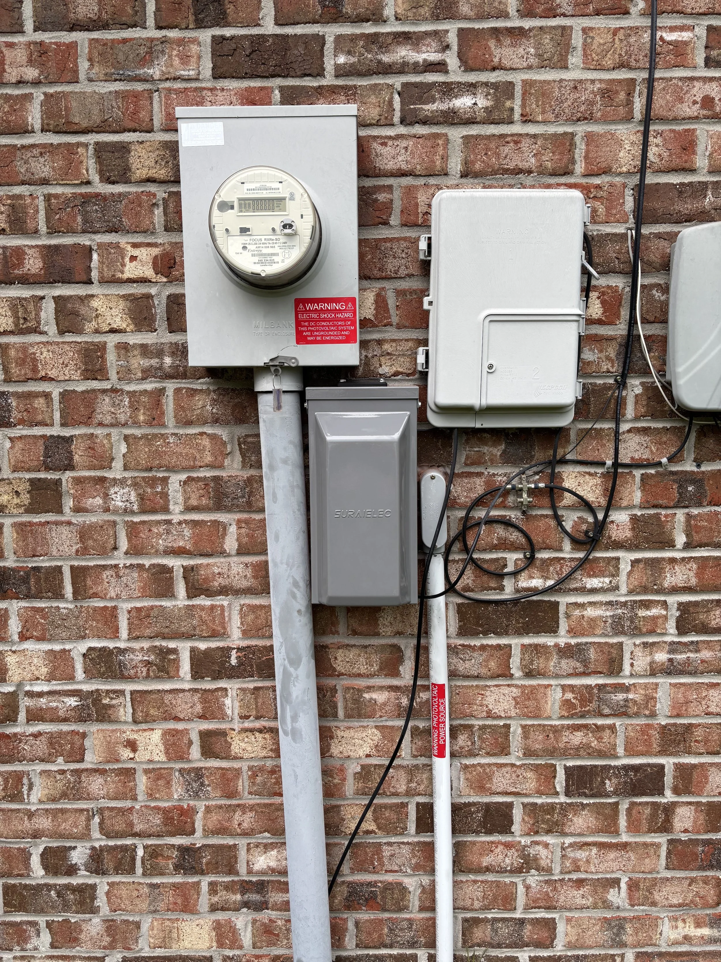 Electric meter, gray electrical box, and wiring mounted on a brick wall.