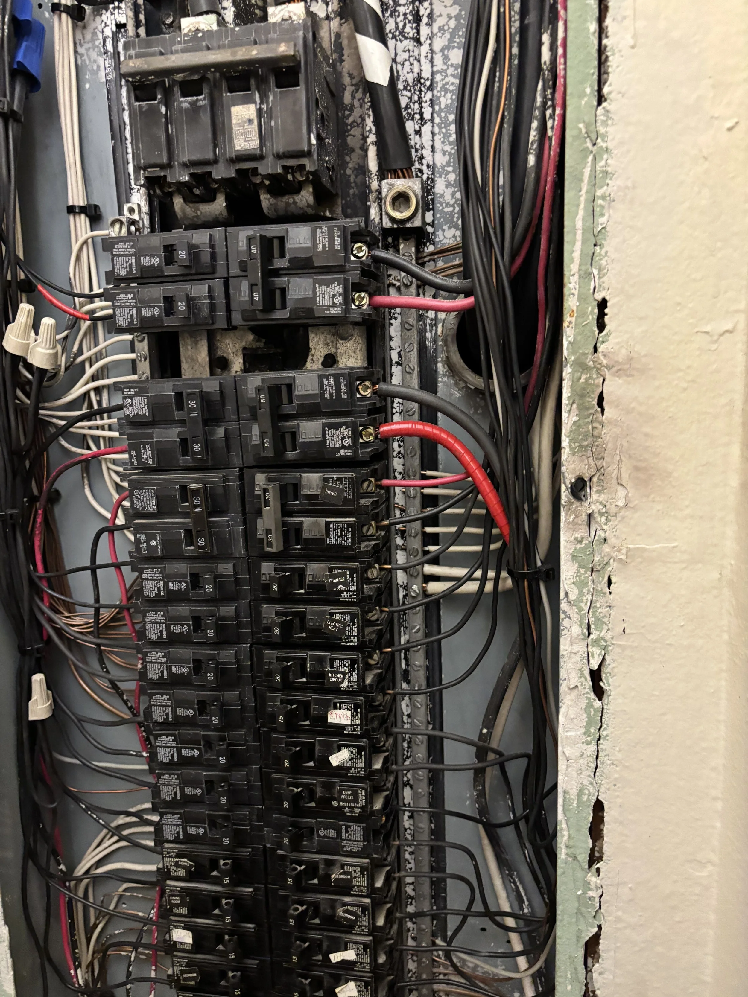 Electrical panel with multiple circuit breakers, wires, and some exposed wiring on the wall.