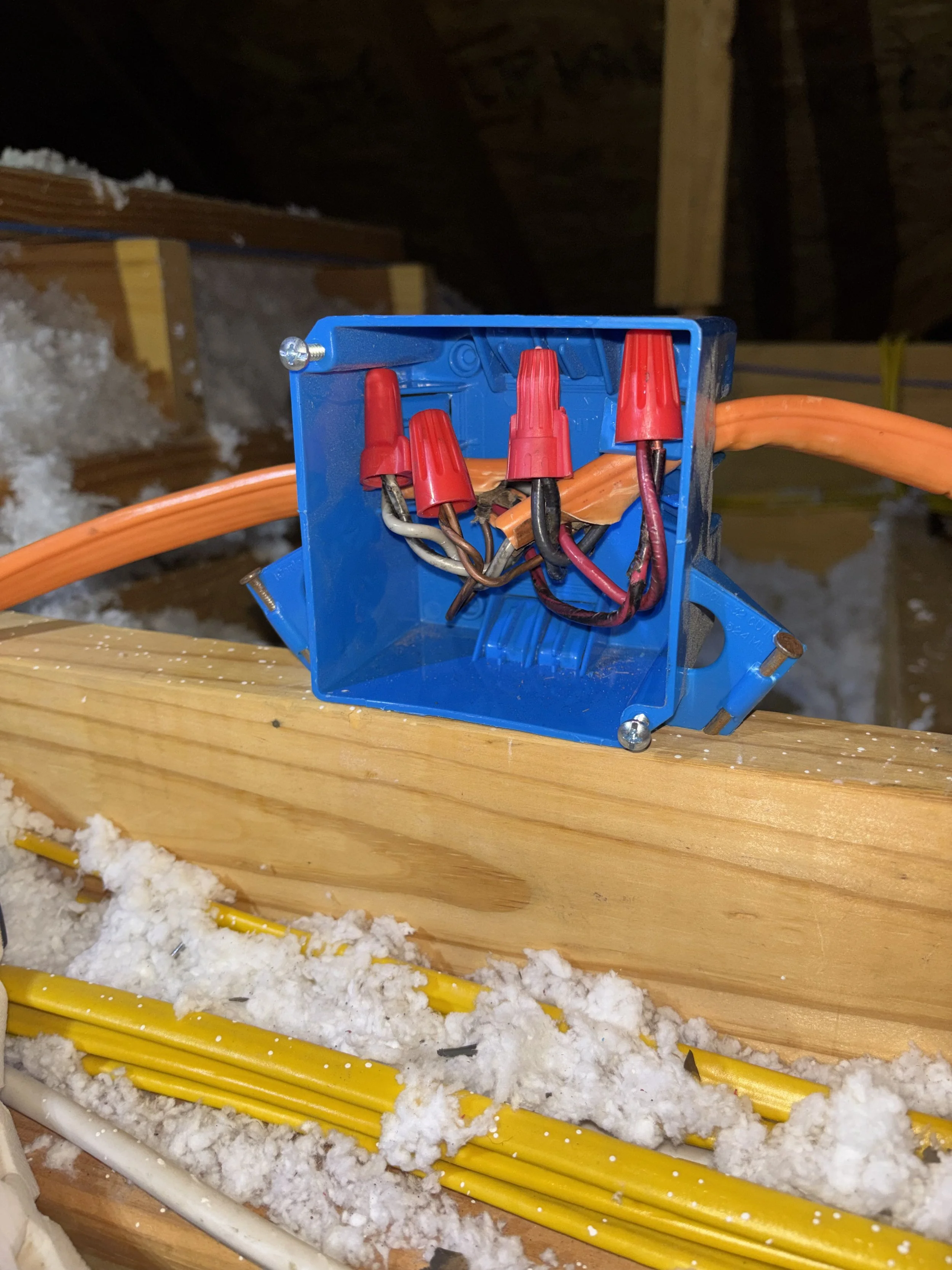 Electrical junction box with red wire nuts, orange electrical wires, and a blue box mounted on a wooden beam in an attic space. Insulation and additional wiring are visible below.