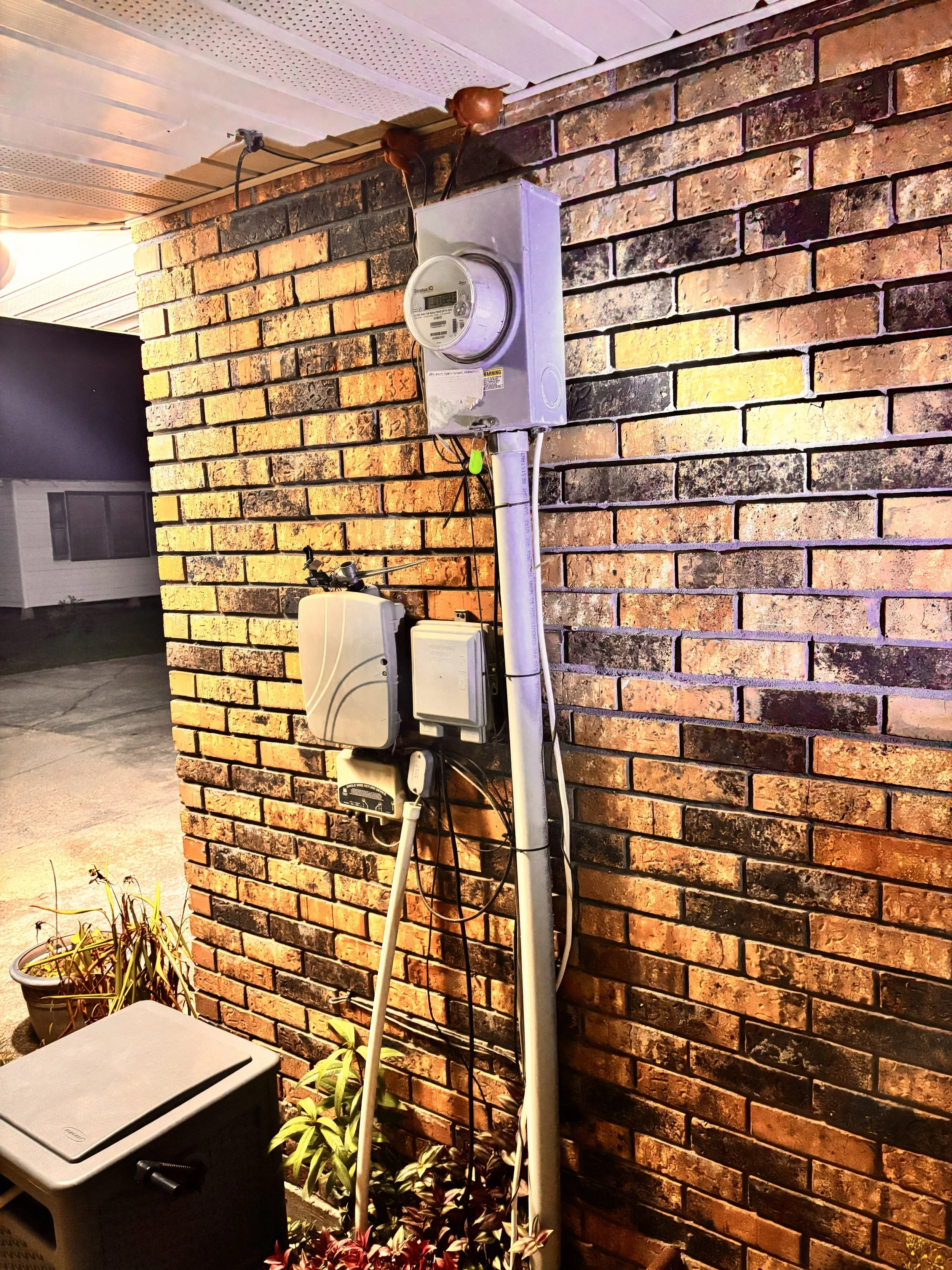 Exterior view of a brick wall with electrical meters, transformers, and wiring mounted on it, near some potted plants and a utility box at night.