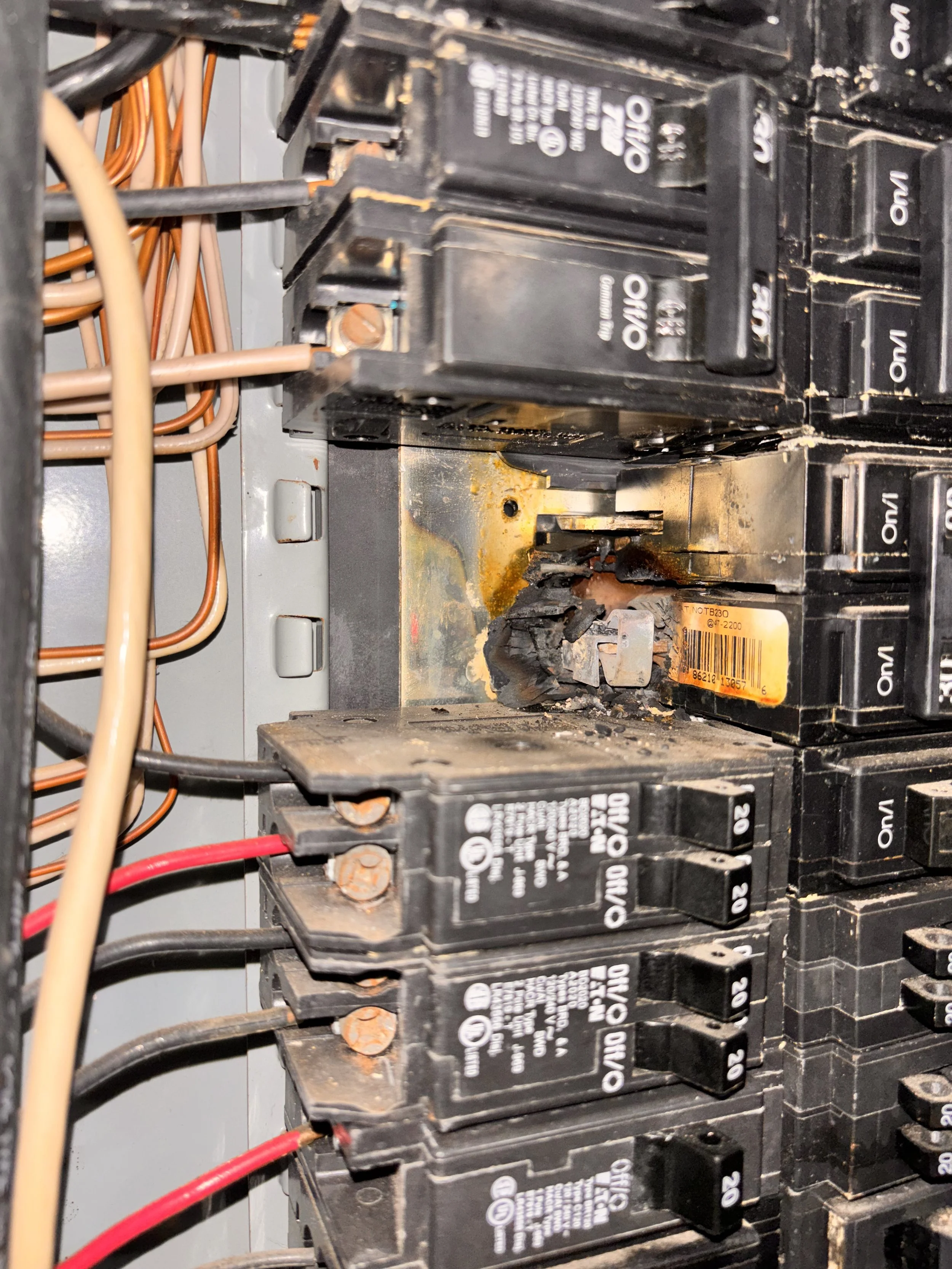 An electrical panel with several circuit breakers, one of which is damaged and burned, with charred debris and rust around it.