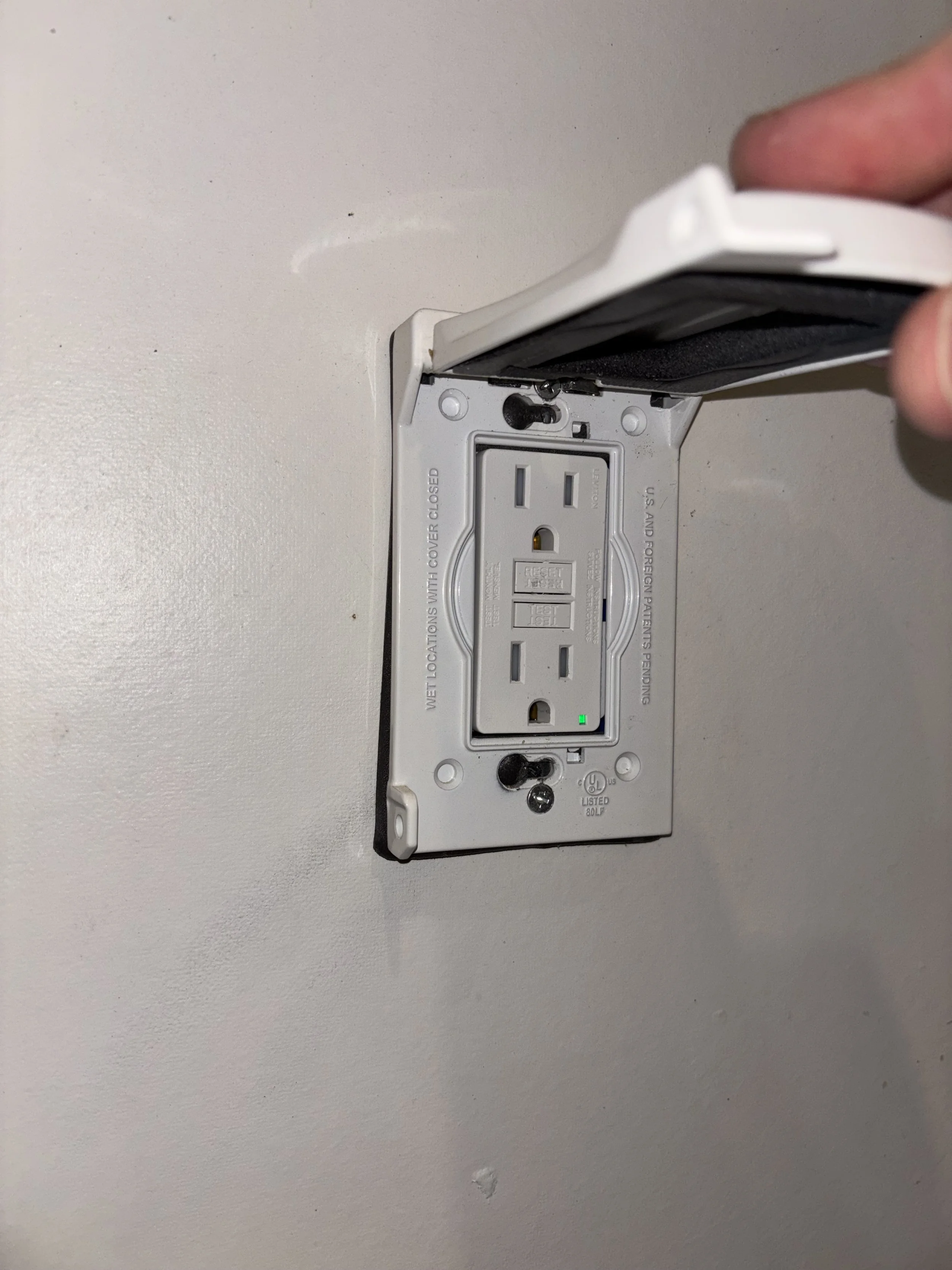 Close-up of an electrical outlet with a black cover being lifted, revealing the wiring and green indicator light.
