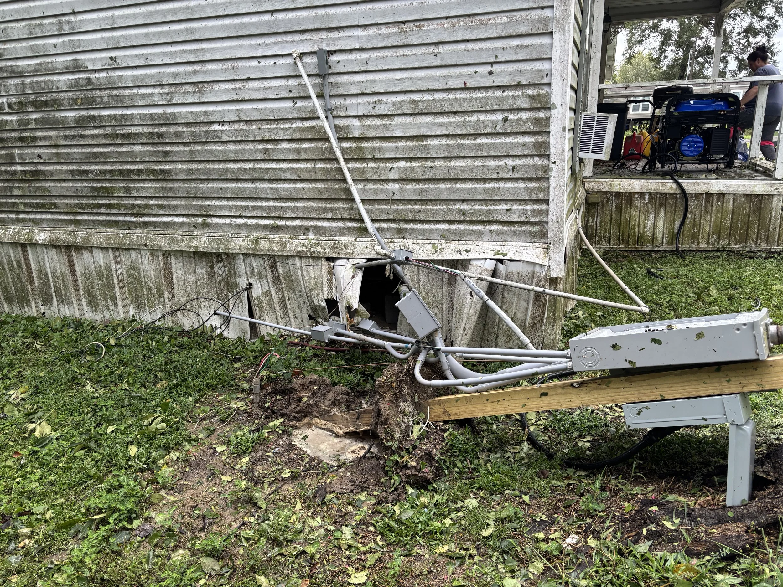 Detached electrical pole and wires fallen against the side of a house, with electrical boxes and cables on the ground, indicating damage from a storm or accident.