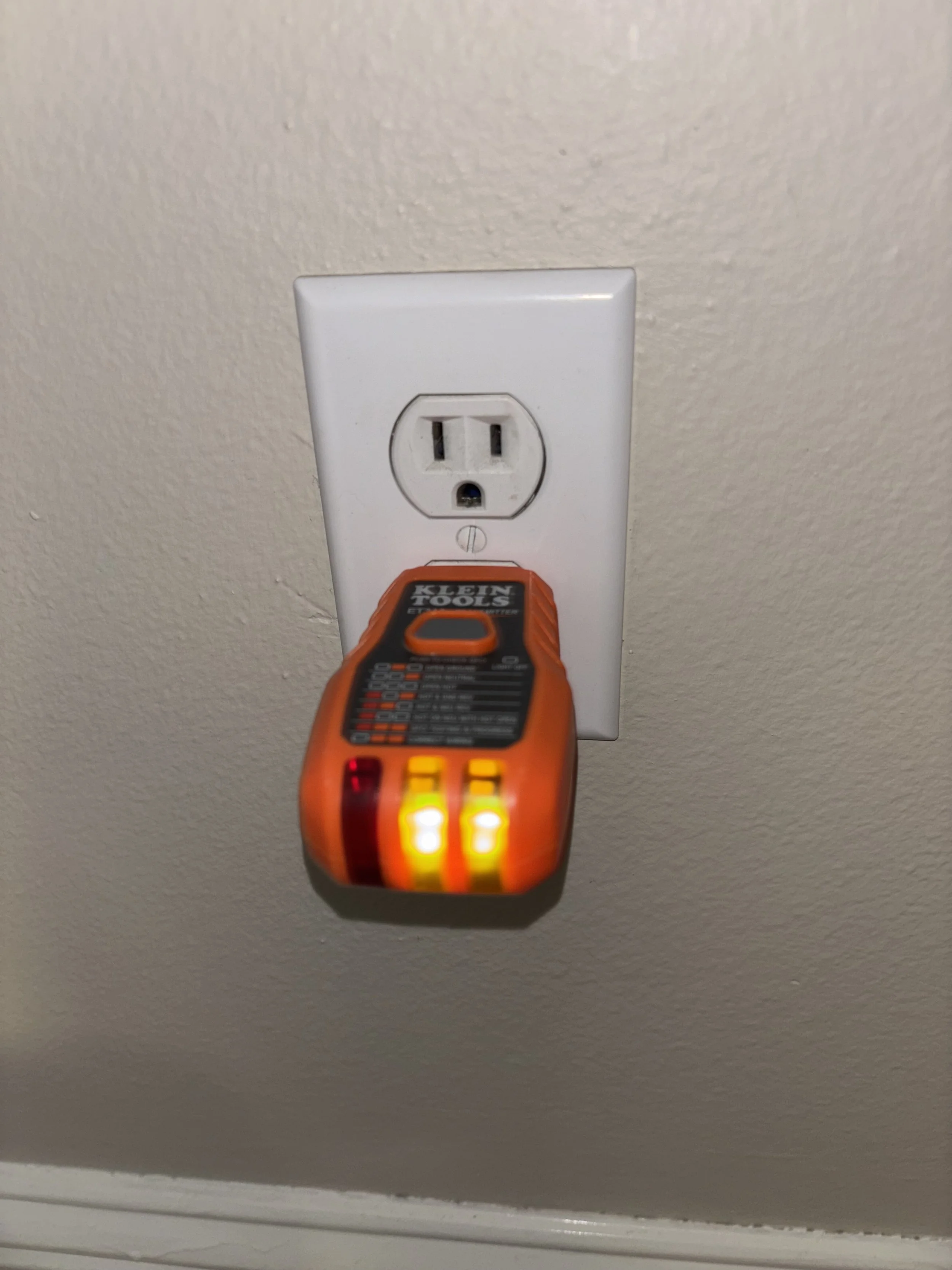 A voltage tester with orange body and yellow and red lights is plugged into an electrical outlet on a wall.