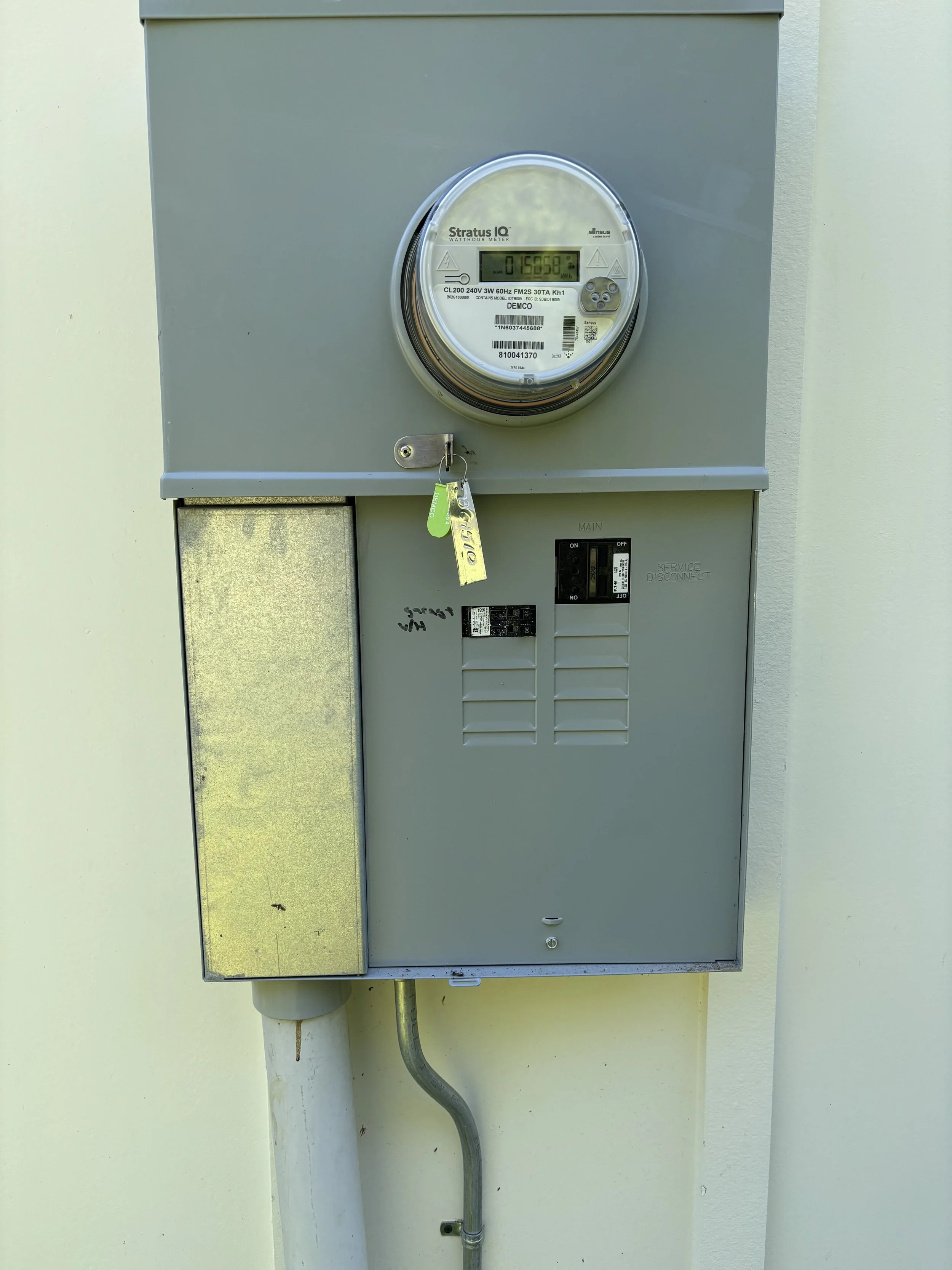 An electric utility meter mounted on a gray metal box on a wall, with a key hanging from it, and electrical wiring below.