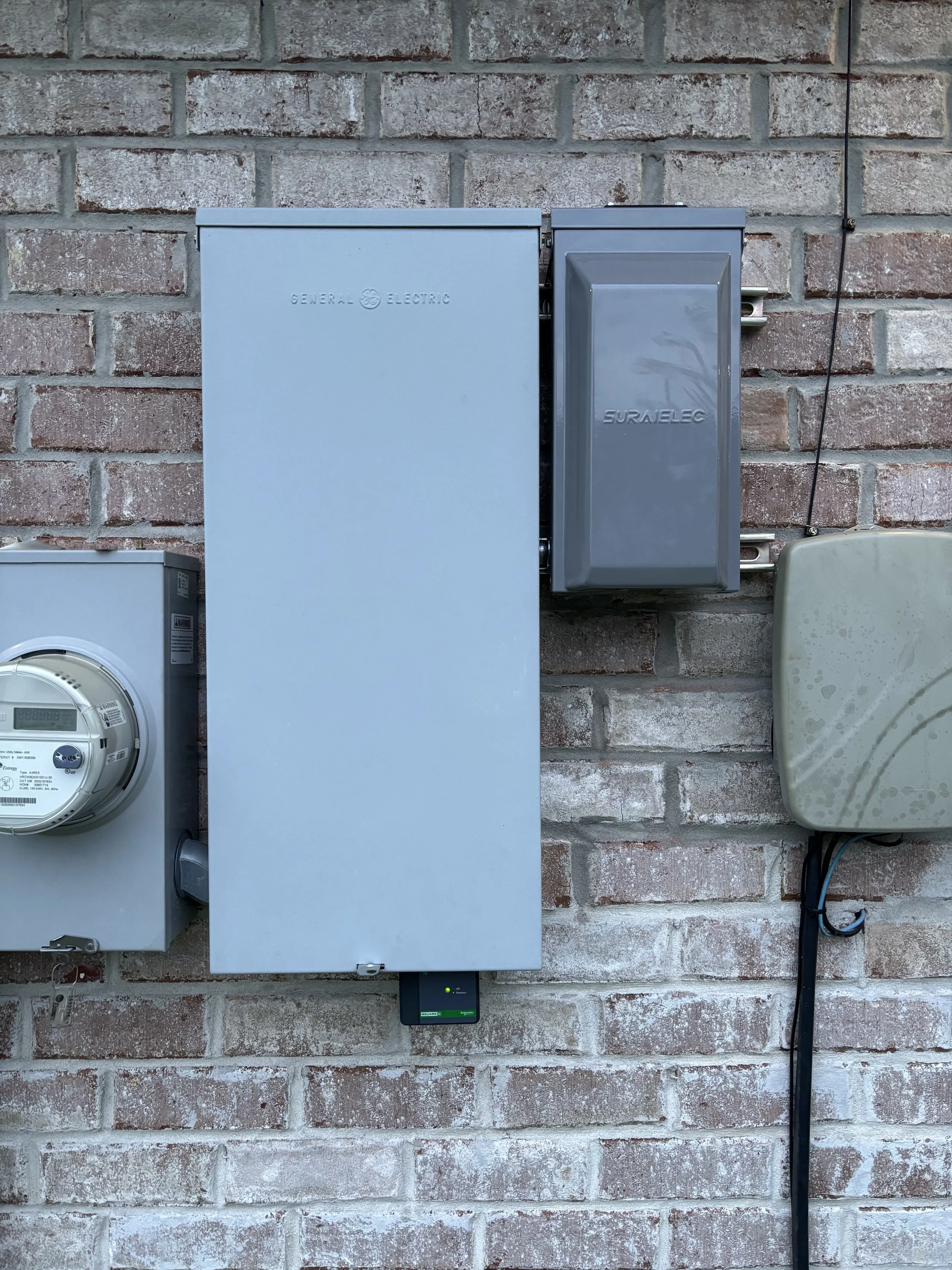 Electrical meters and boxes mounted on a brick wall, including a gray electric meter on the left, a large light gray electrical box in the center, and a dark gray box labeled Suralelec on the right.