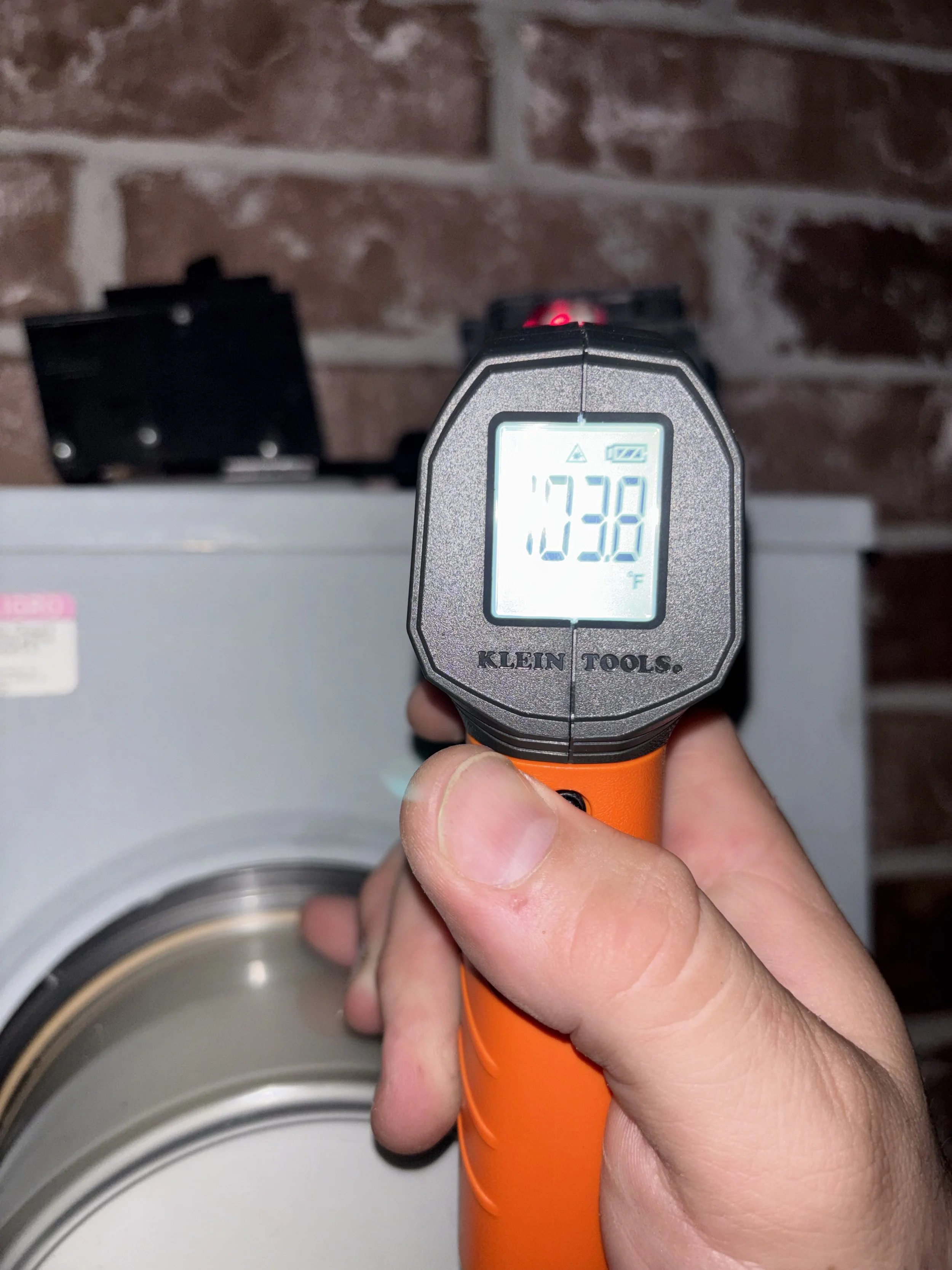 A person's hand holding a portable infrared thermometer aimed at a washing machine, displaying a temperature reading of 103.8 degrees Fahrenheit.