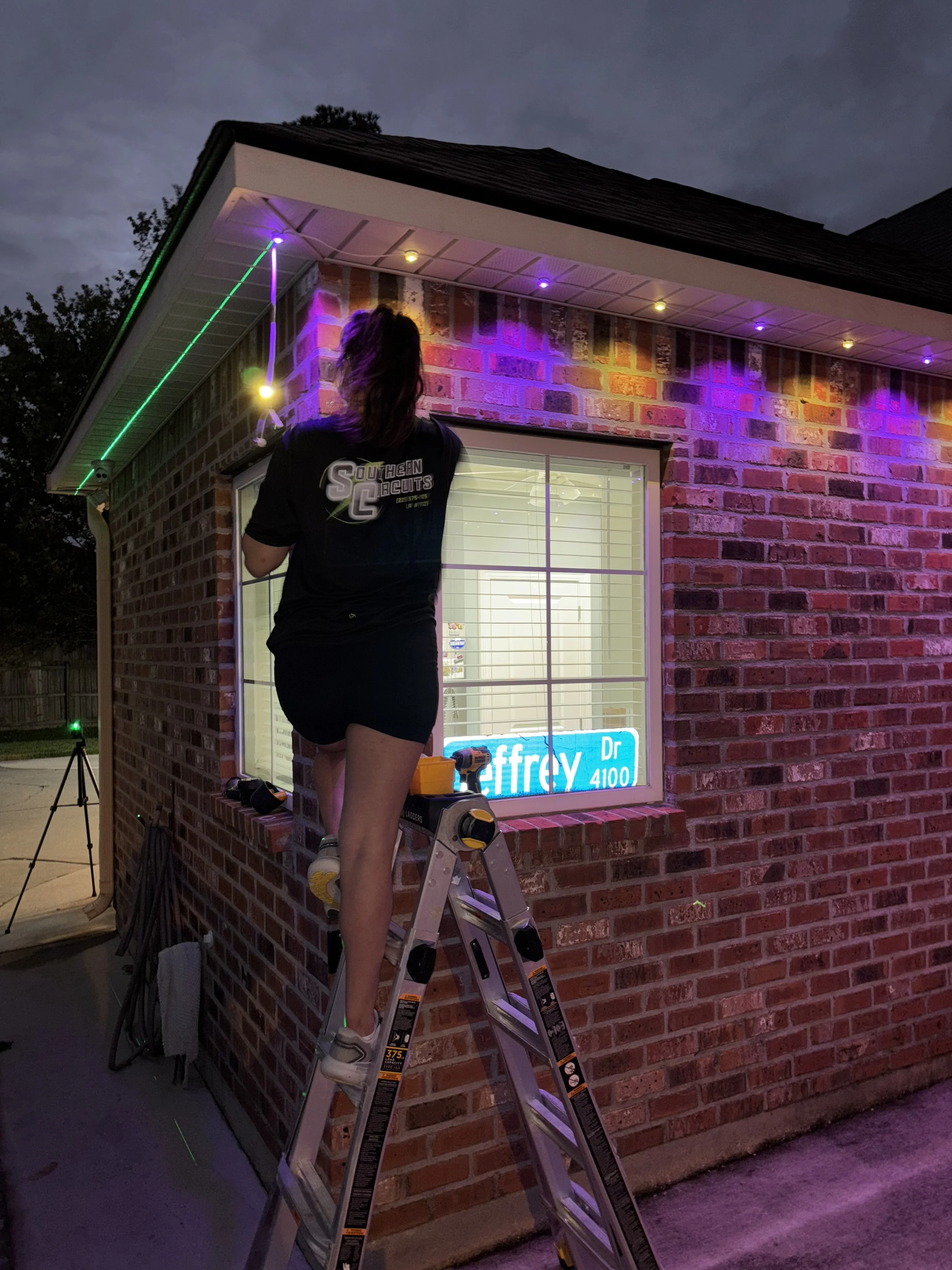 Woman on a ladder installing colorful LED lights on a brick house at dusk, with a window displaying a neon sign inside.