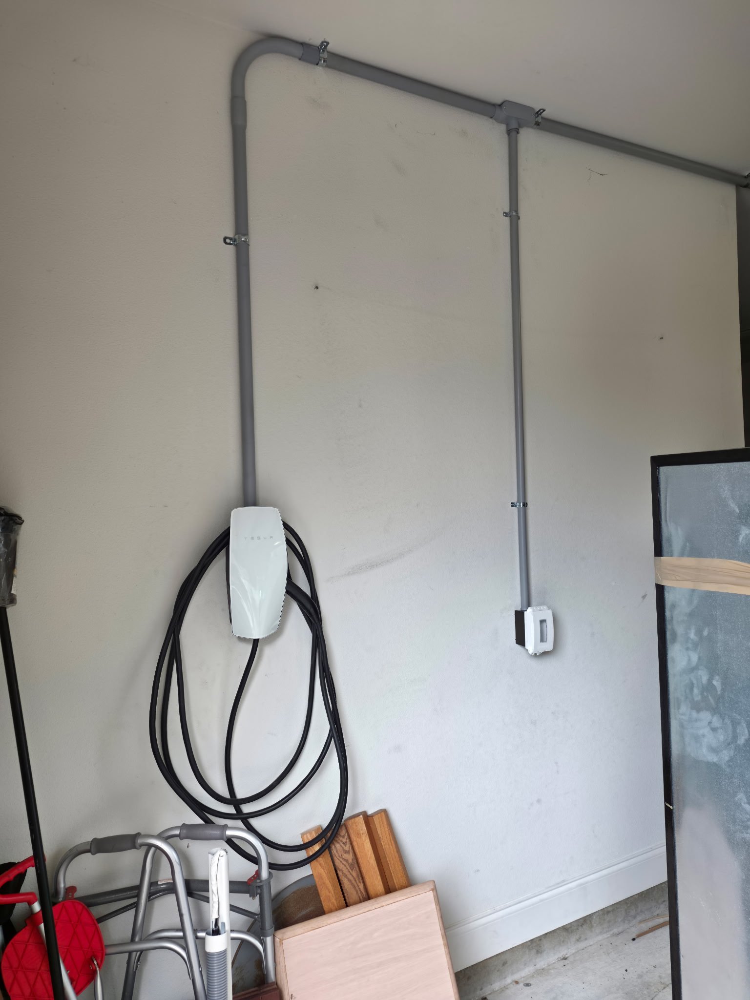 Electric vehicle charging station and electrical conduit pipes installed on a white wall in a garage, with some wooden objects and tools on the floor.