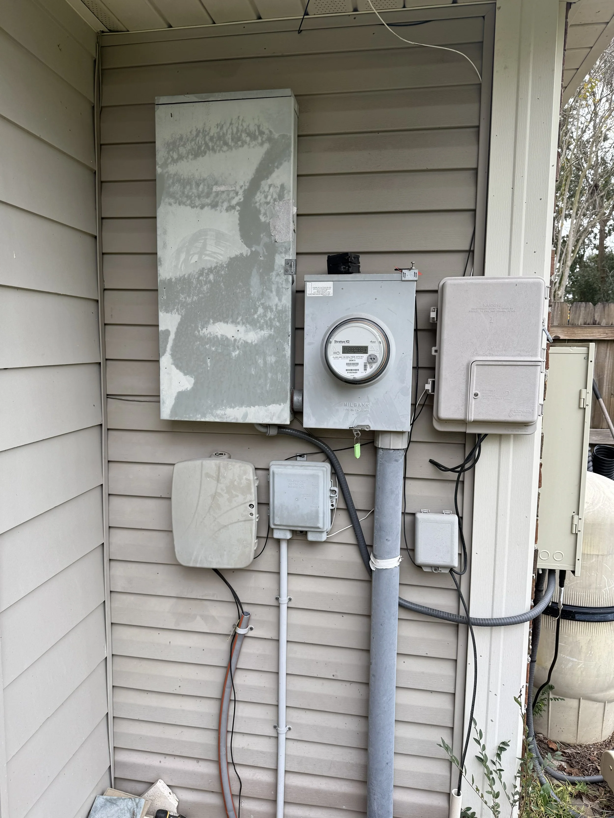 Outdoor utility area with electrical meters, panels, and conduits mounted on house siding.