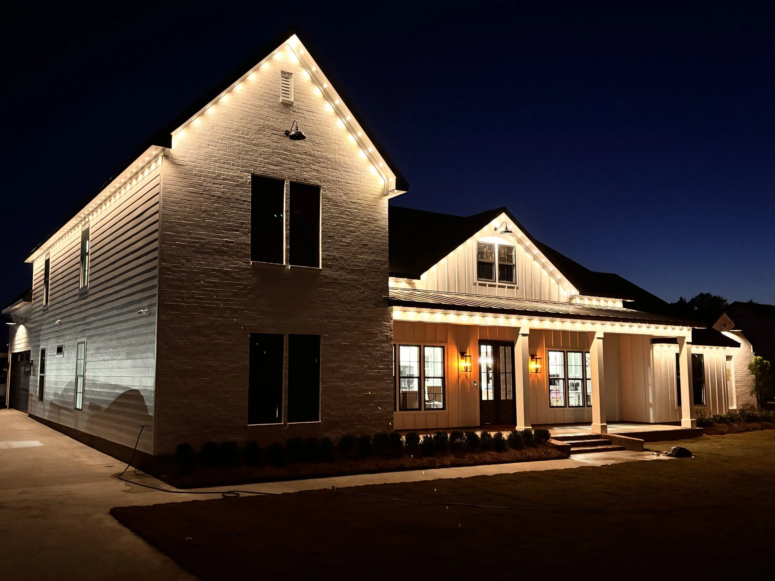 A house decorated with string lights at night, featuring a brick and siding exterior, porch lights, and a neatly maintained lawn.