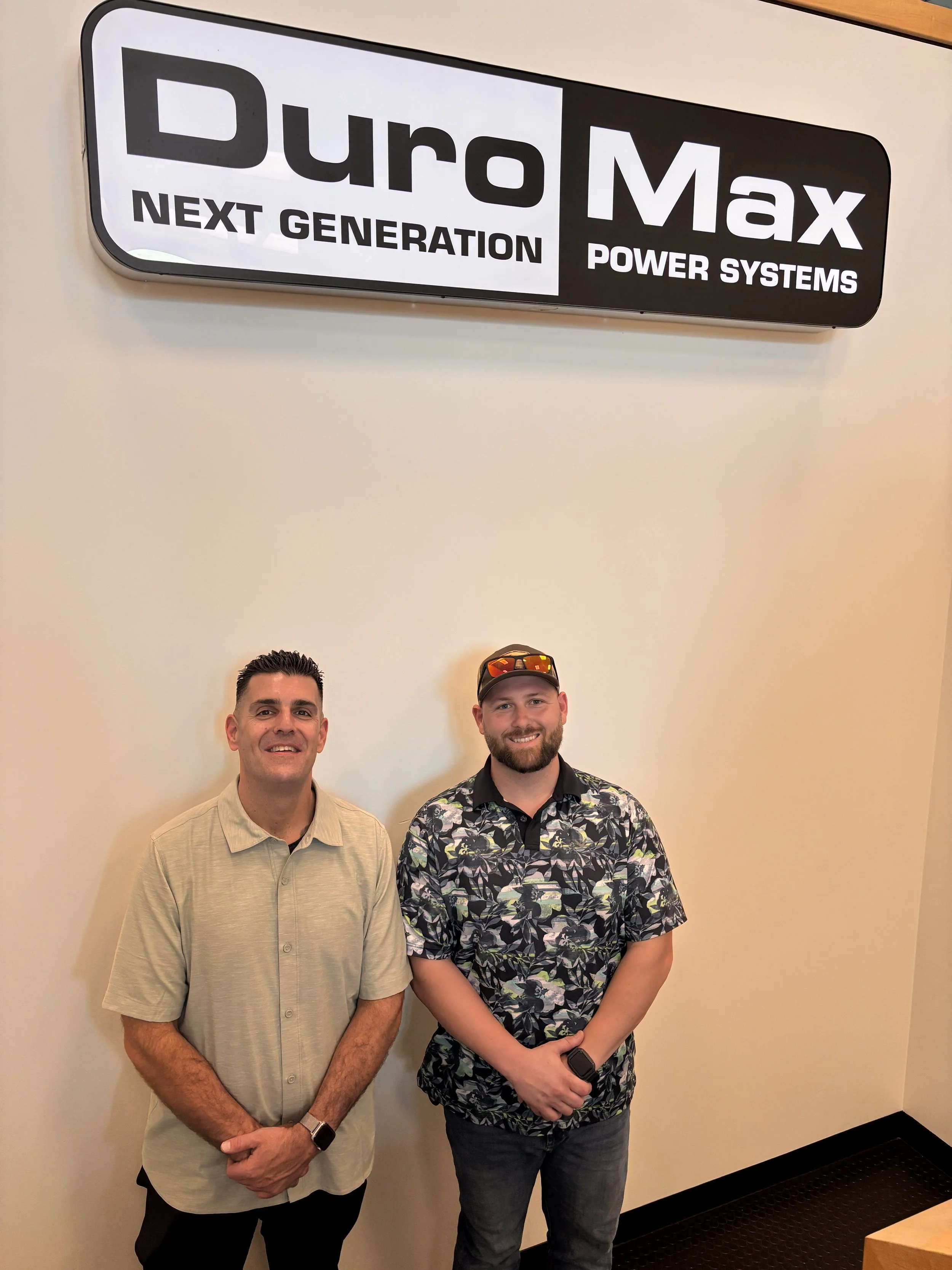 Two men standing indoors, smiling, in front of a wall with a sign that reads "Duro Max Next Generation Power Systems."