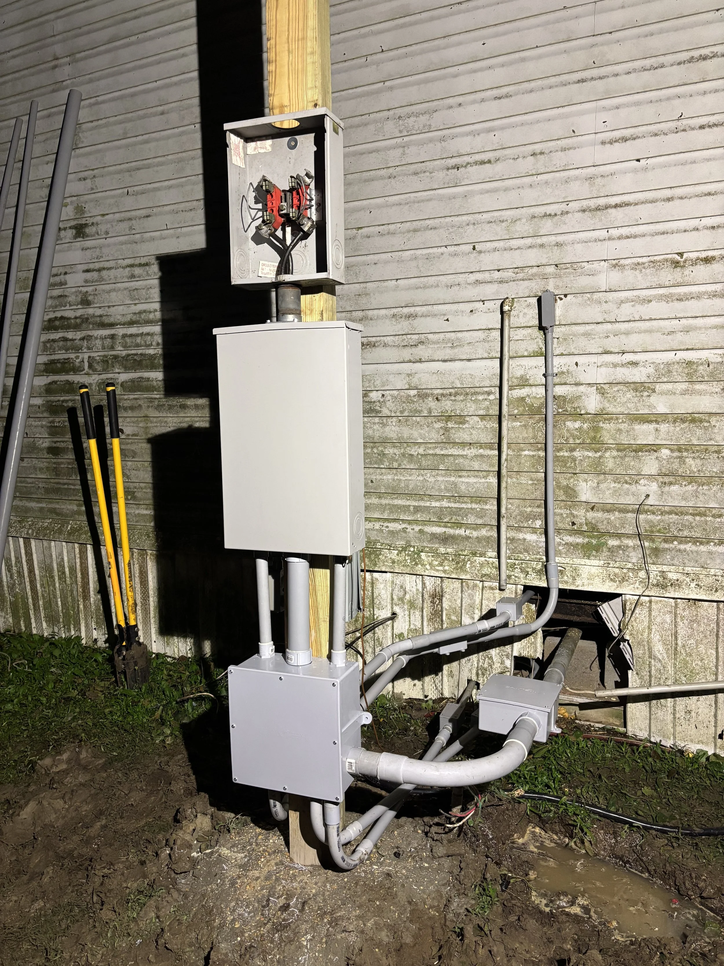 Electrical meter, junction box, and conduit pipes attached to a wooden pole and the side of a building.