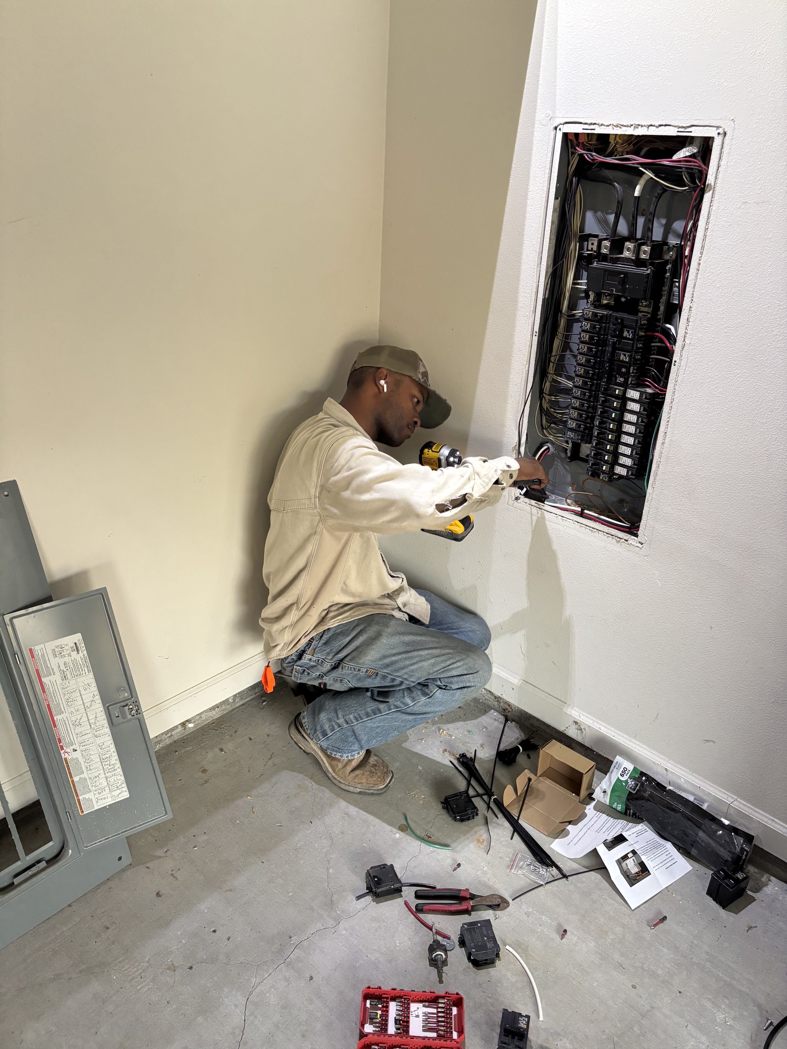 A technician installing or repairing an electrical panel in a wall. He is crouched down with tools and parts on the floor around him, working on the circuit breaker box.