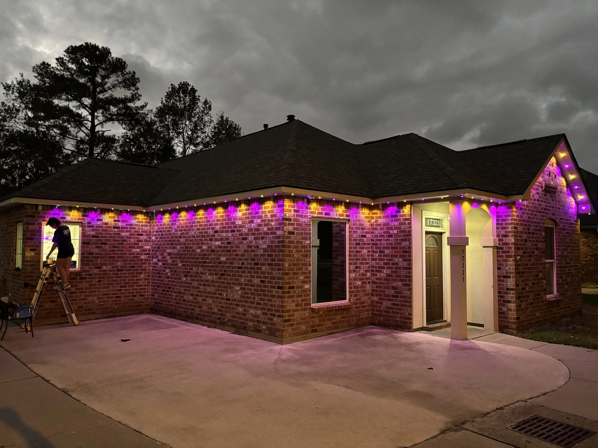 A brick house with purple and yellow Christmas lights along the roofline, a person on a ladder adjusting lights, and a cloudy evening sky.