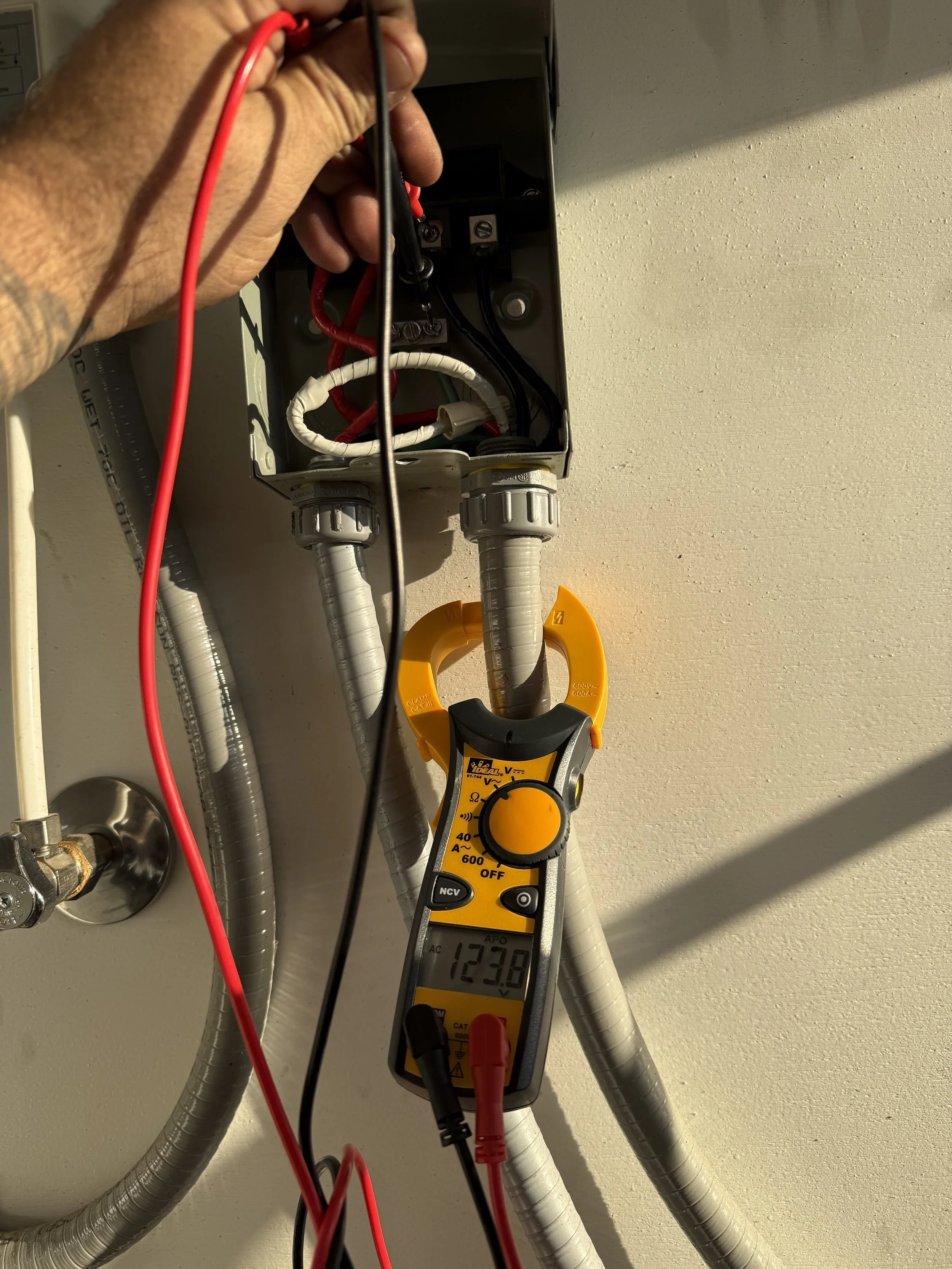 A person using a digital multimeter to measure voltage from an electrical box, with red and black probes connected to the device, and wires inside the electrical box.