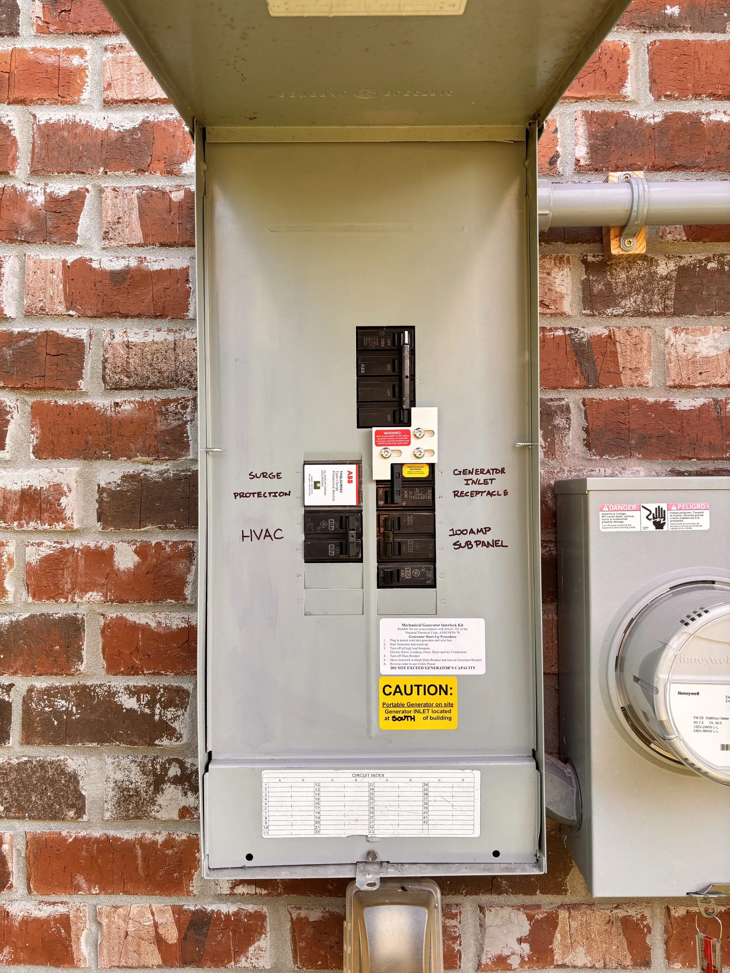 Electrical panel box mounted on a brick wall, with labels indicating surge protection, HVAC, generator inlet receptacle, and 100AMP subpanel, along with a caution sticker and circuit index chart.