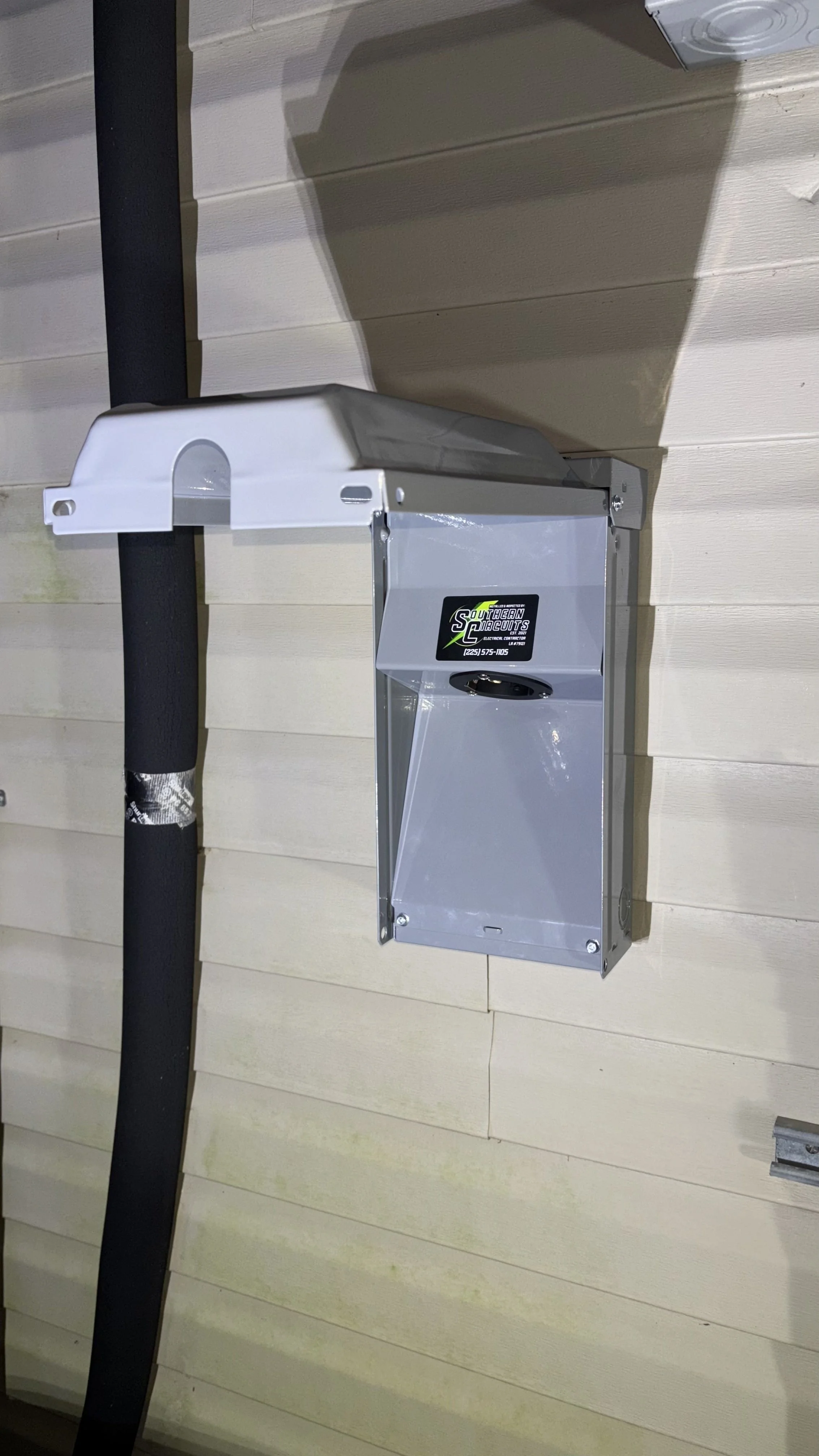 A gray metal electrical box mounted on a beige wooden exterior wall of a building, with a black insulated pipe running vertically beside it.