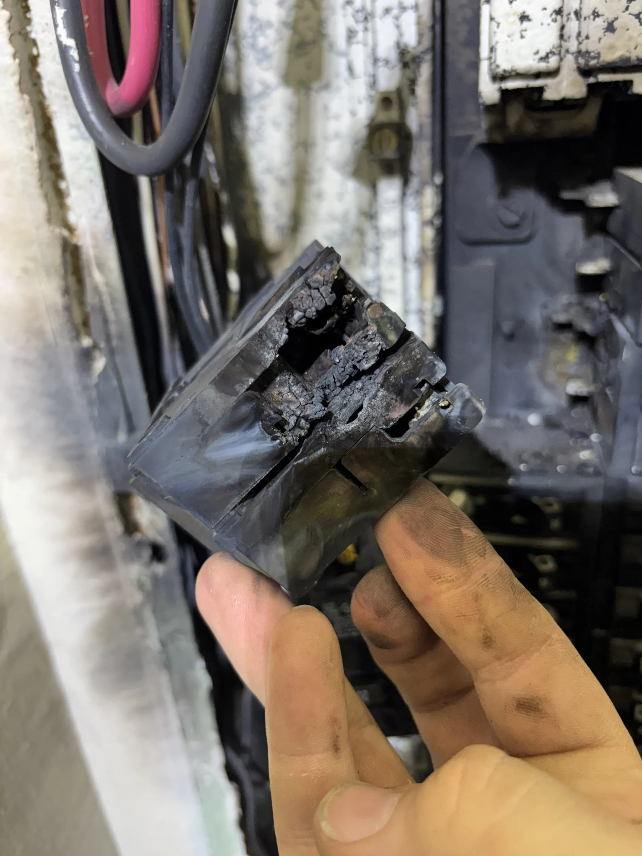 Close-up of a burnt electrical component held by a person's fingers, with visible damage and melting, in a mechanical or electrical setting.