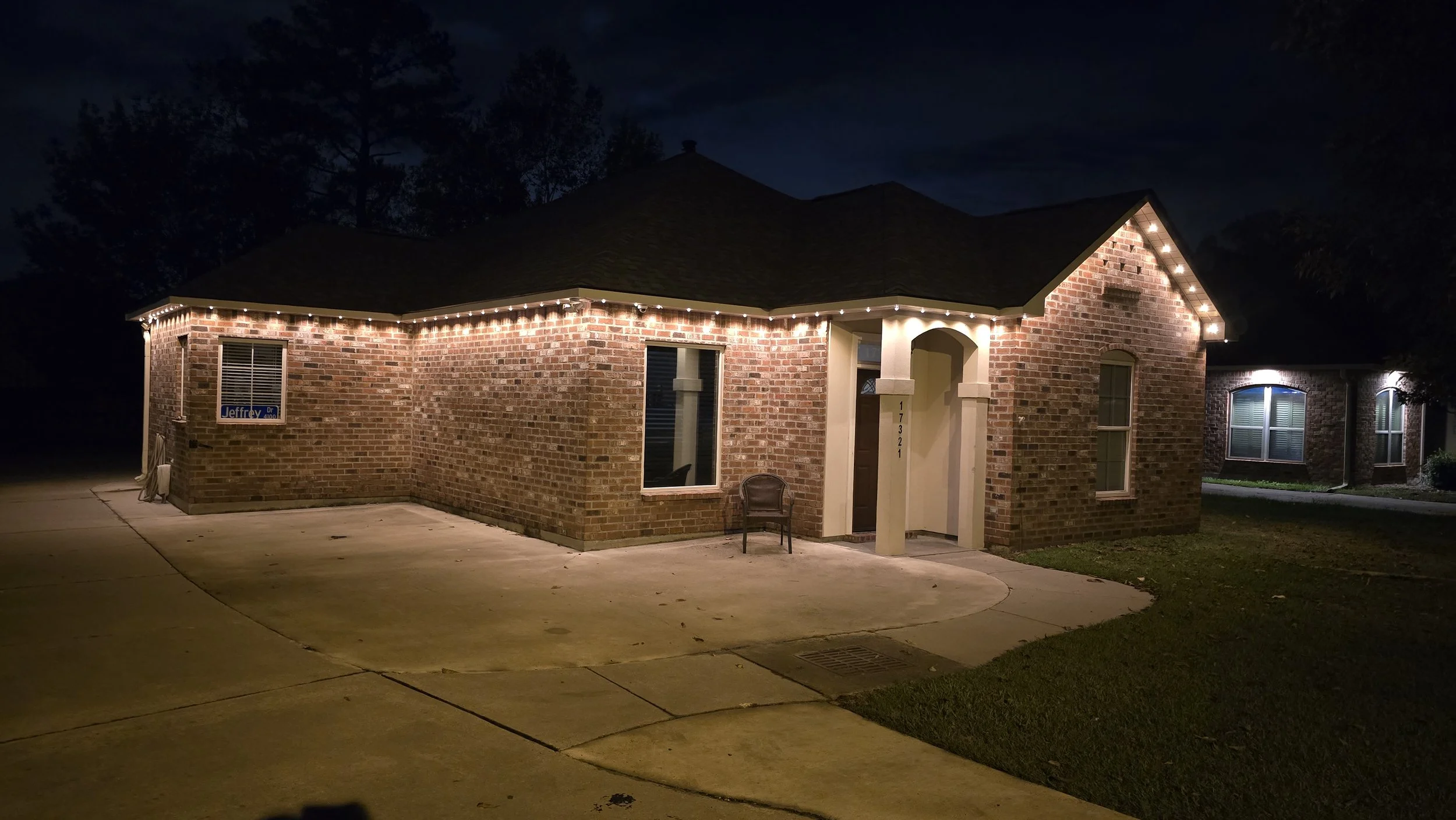 A brick house decorated with white string lights along the roofline at night. There are two large windows, a small front porch with a black chair, and a driveway in front.