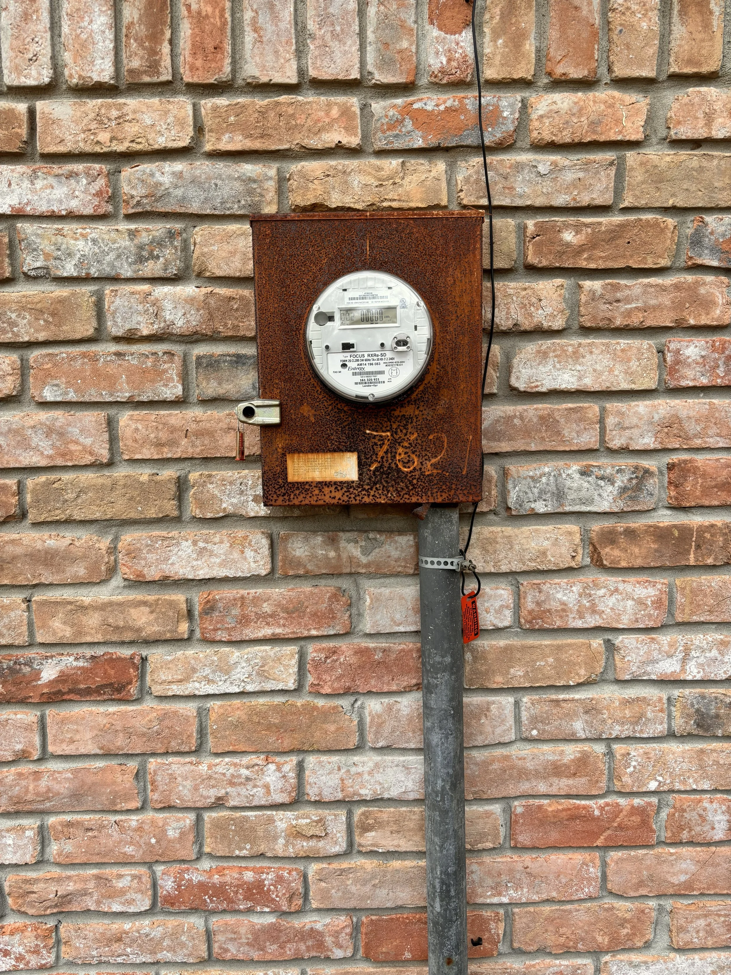 Rusty electrical meter box mounted on a brick wall with a black wire hanging down.