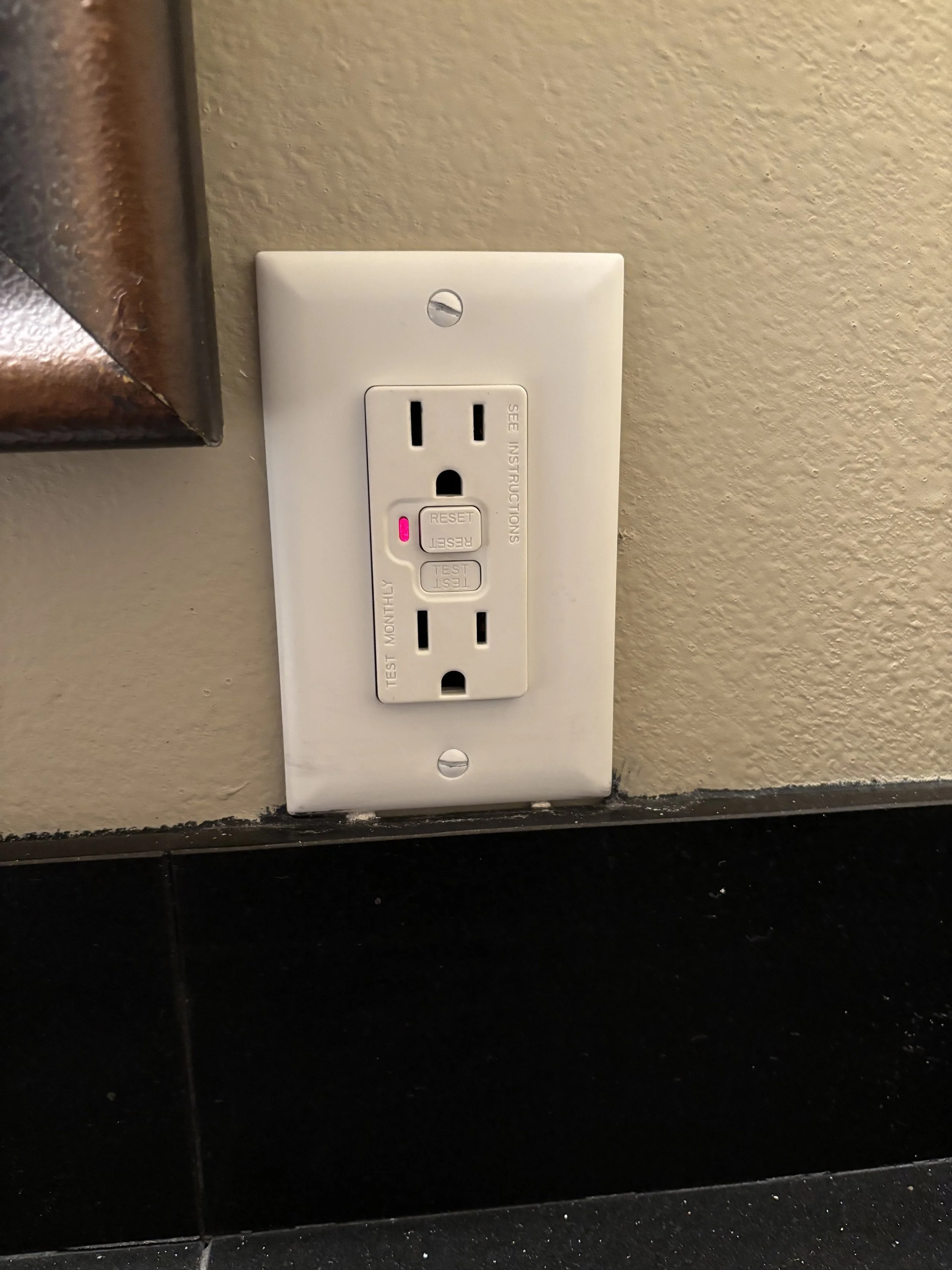 Electrical outlet with a built-in tester and reset buttons, mounted on a wall near a corner with black flooring beneath.