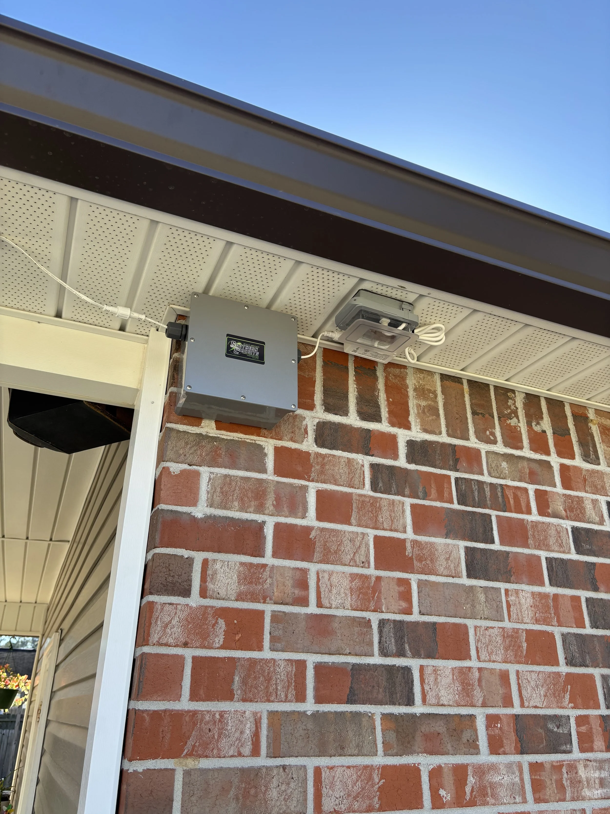 Close-up view of the corner of a house showing a brick wall, part of the roof, a gray electrical box, and an outdoor light fixture under the soffit.