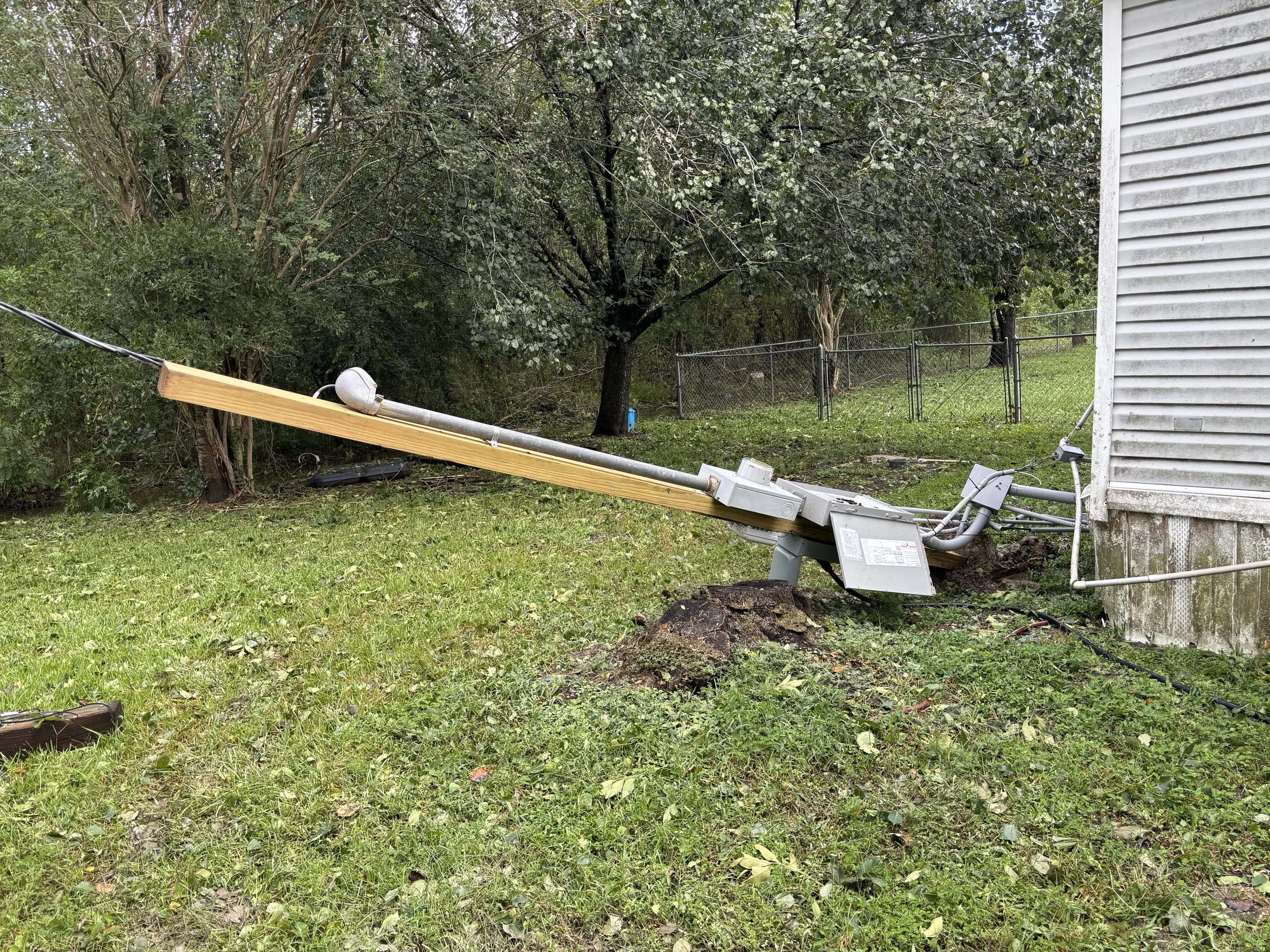 Damaged utility pole leaning against a house with wires disconnected, in a grassy backyard with trees and a chain-link fence.