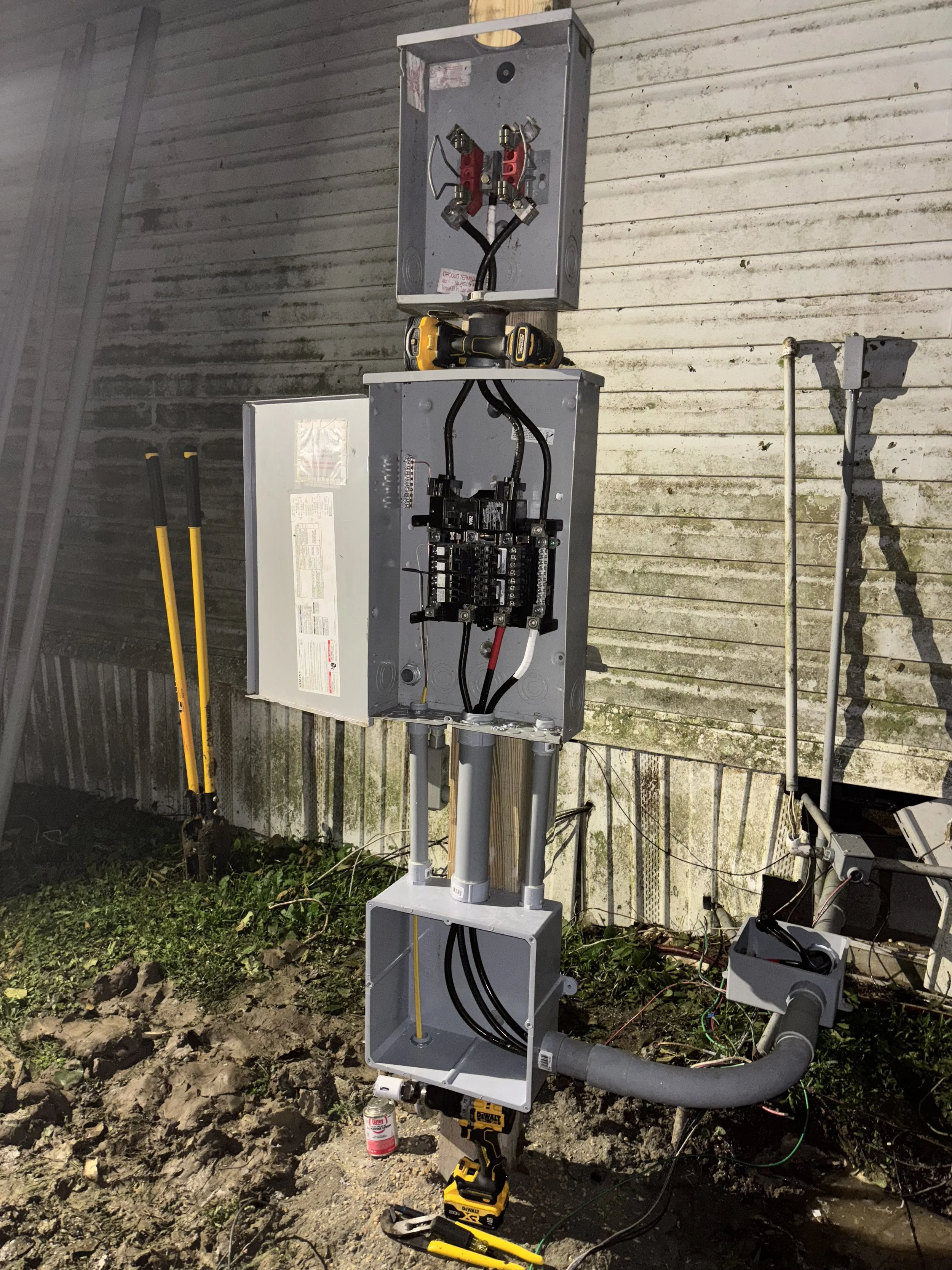 Outdoor electrical panel and conduit box being installed, with power tools and wires nearby, against a weathered wooden wall.