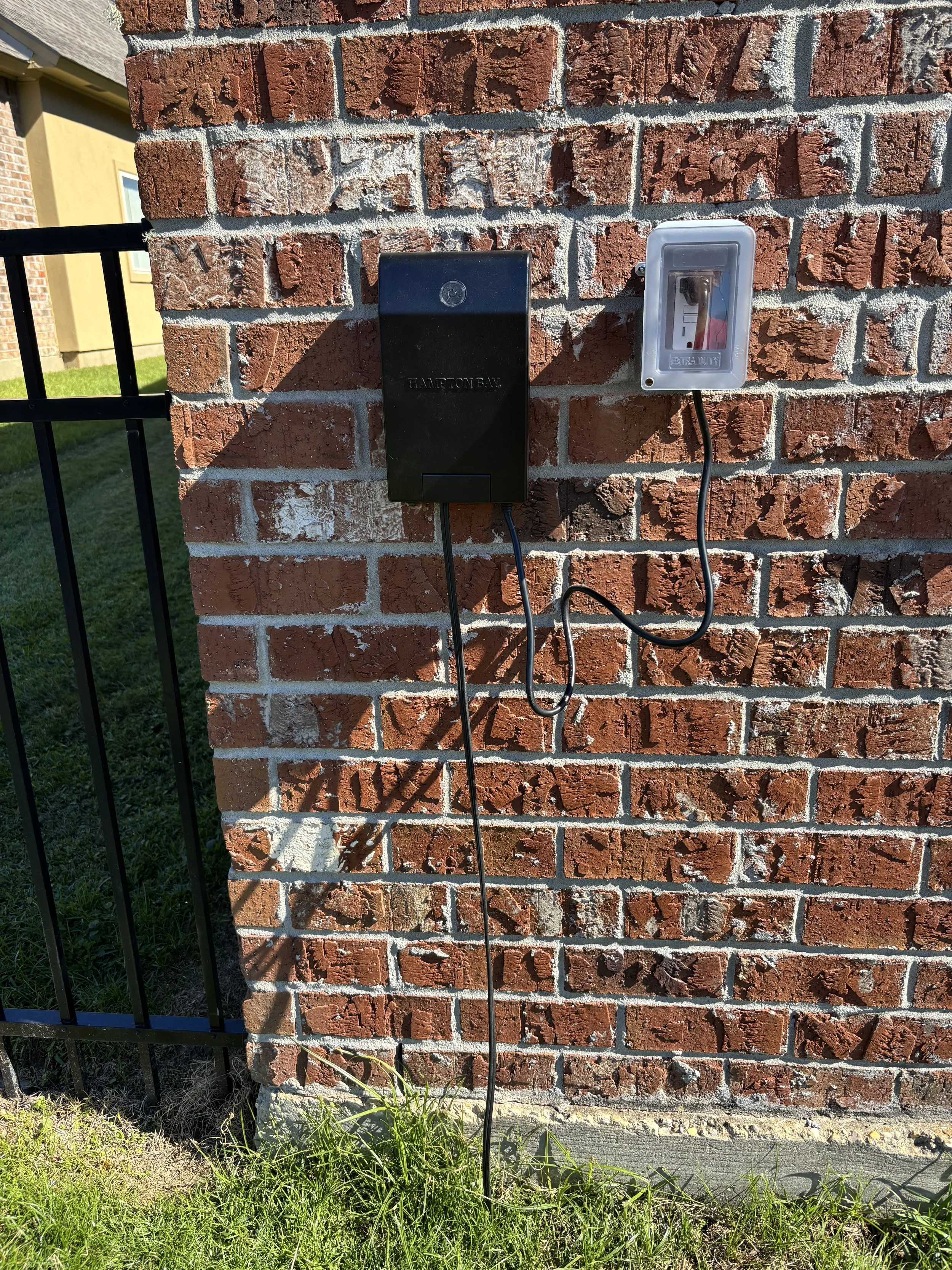 Electric vehicle charging station mounted on a brick wall near a black metal fence, with a charging cable hanging down and grass at the base.