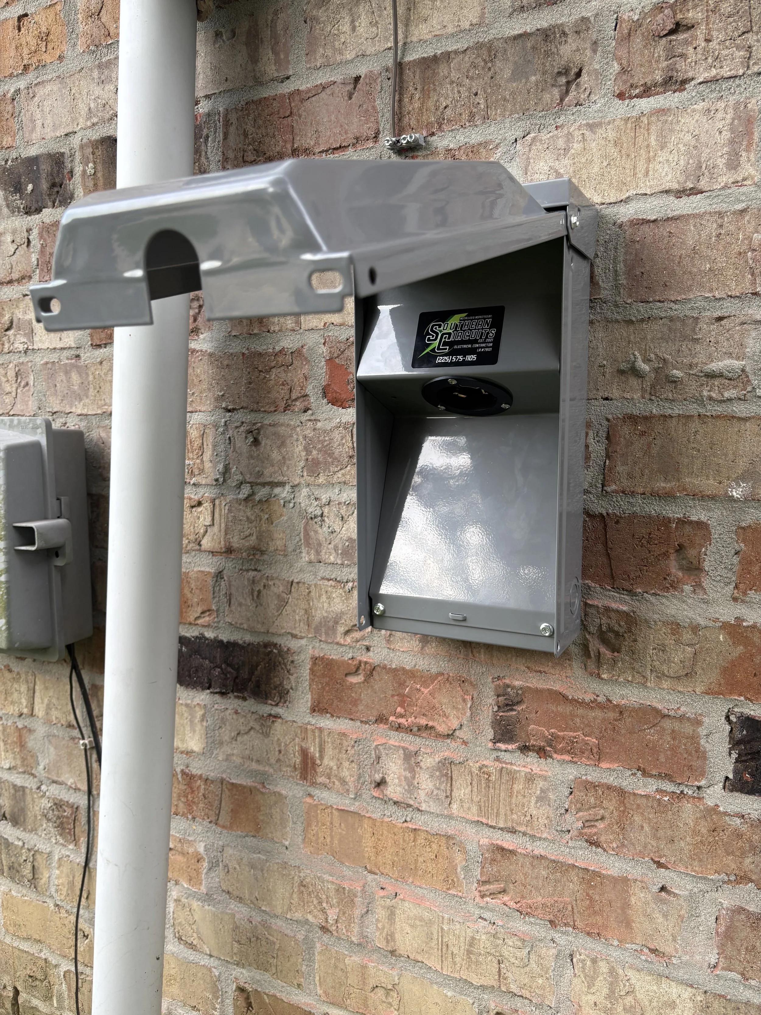 An outdoor electric vehicle charging station mounted on a brick wall, with a gray metal cover partially opened.