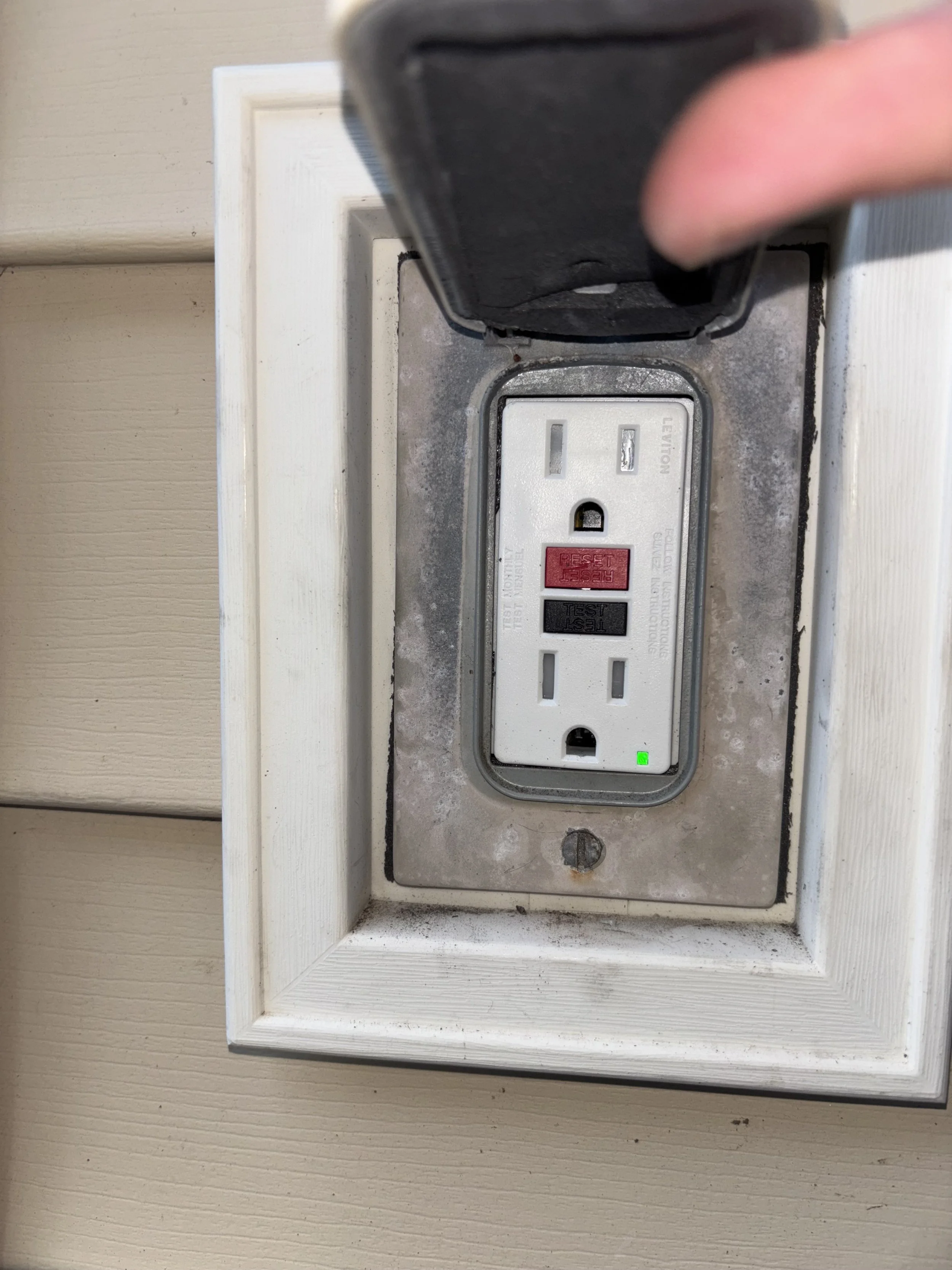 Close-up of a residential electrical outlet with a black cover lid, showing a three-prong socket with a green indicator light, in a white frame, mounted on a beige wall.