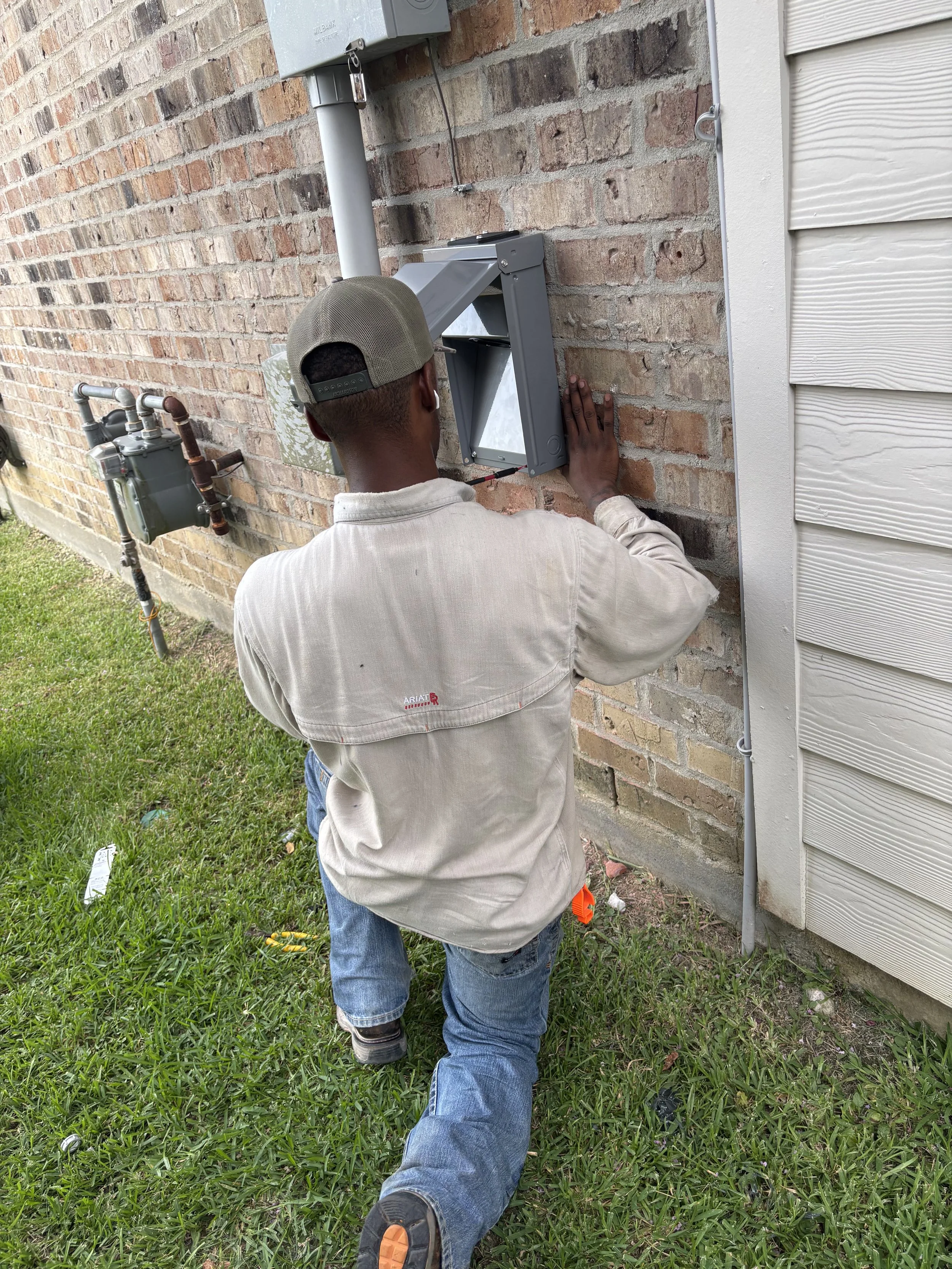 A worker installing or repairing an outdoor utility box on a brick wall outside a house. The worker is wearing a beige shirt, jeans, a backward beige cap, and is kneeling on the grass.