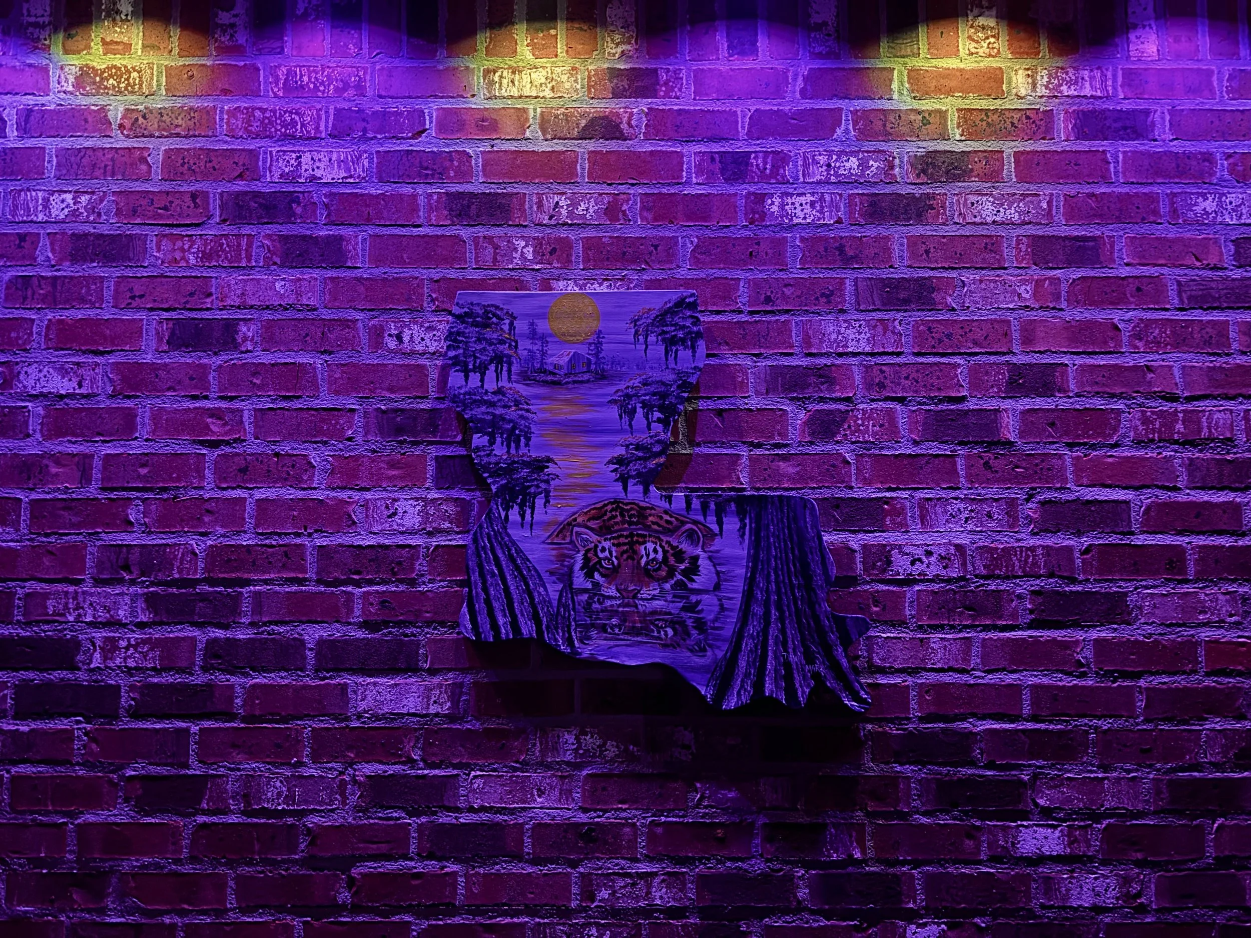 A colorful, artistic painting on a brick wall illuminated by purple and yellow lights. The painting depicts a woman with closed eyes, a tiger's face on her chest, and a scenic background with trees, water, and the sun.