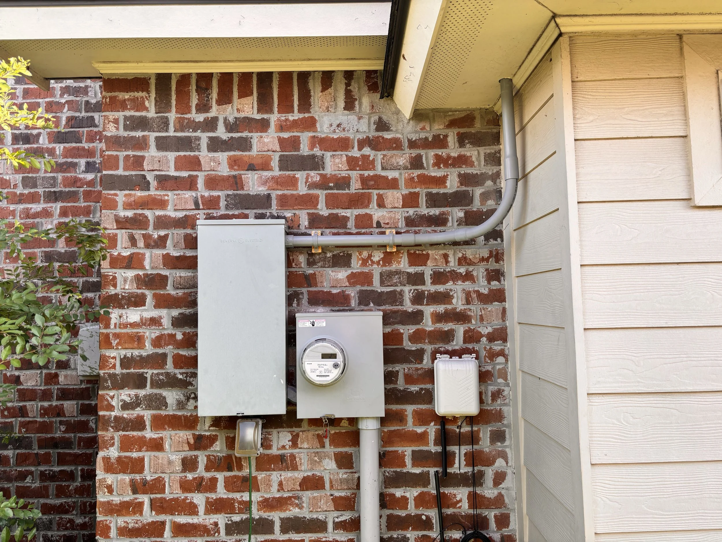 External view of house showing an electrical meter box, a power panel, and cable connections against a brick wall.