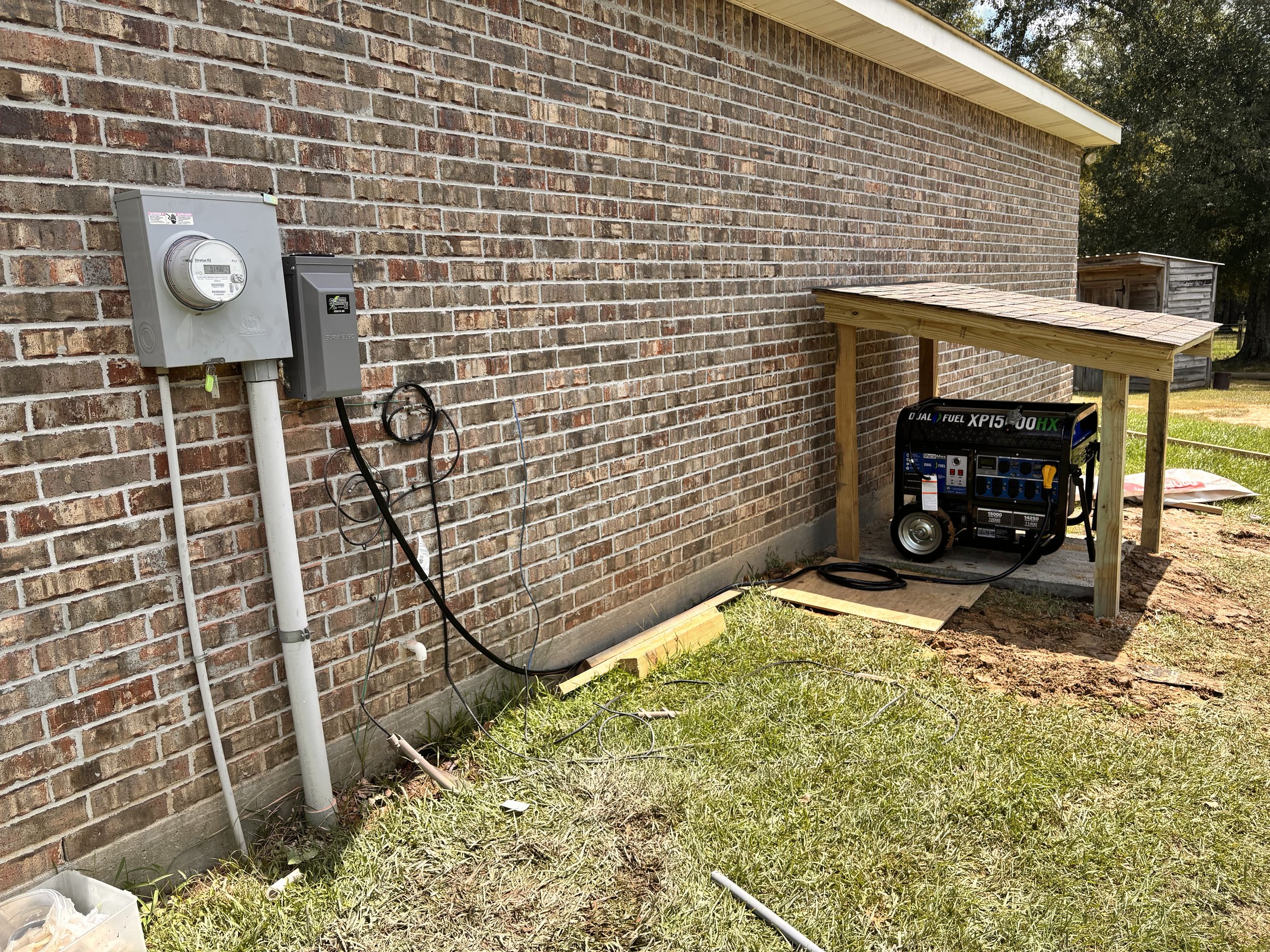 Outside of a brick building with electrical meters, cables, and a portable generator under a small wooden shelter.