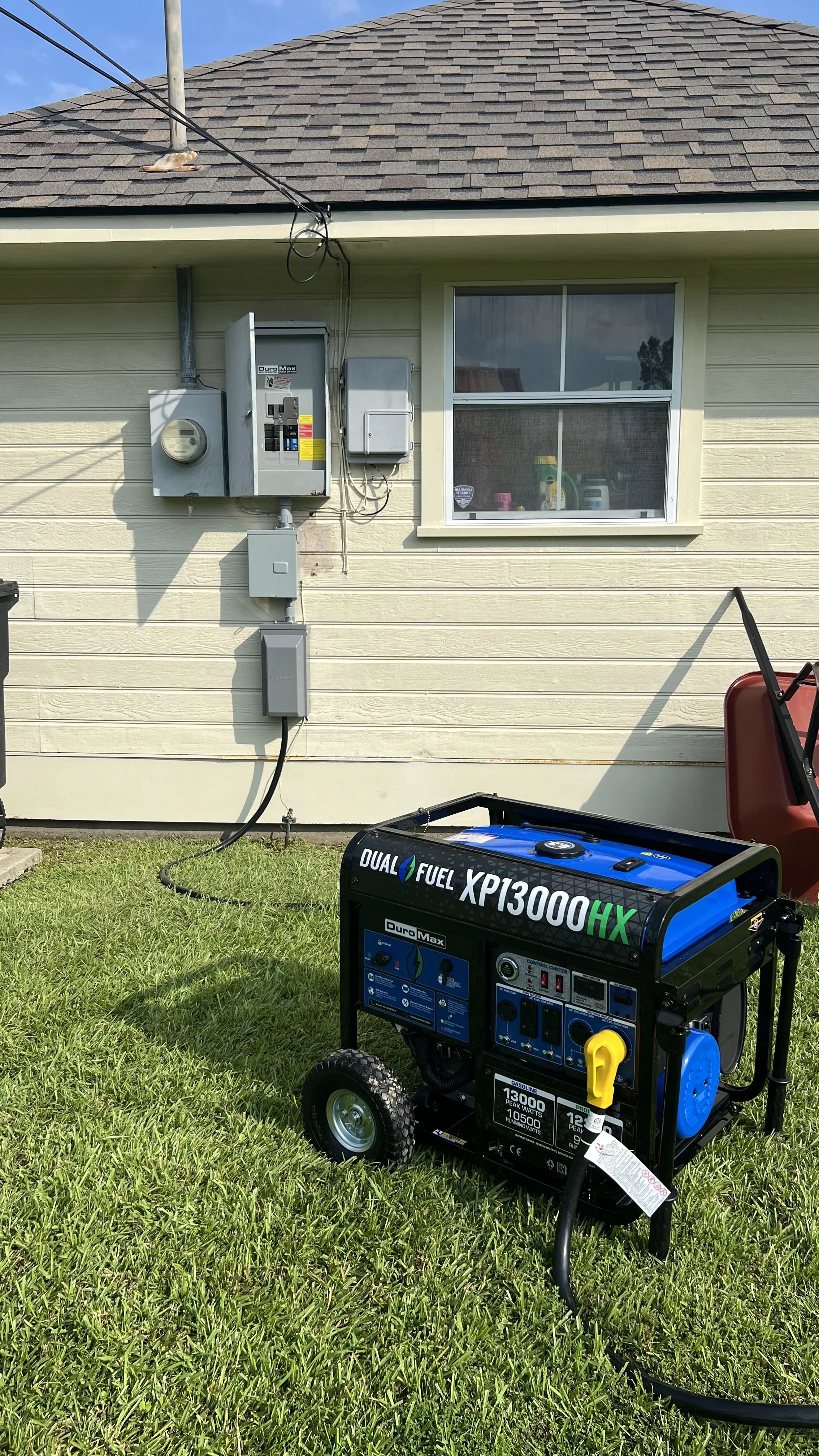 A portable generator on a grassy lawn in front of a house, with electrical panels and a window on the house's exterior wall.
