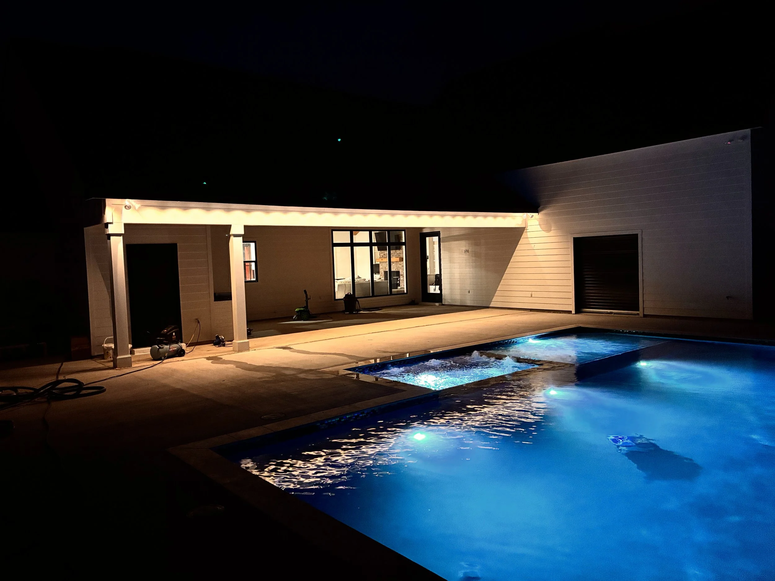 Nighttime view of a backyard with a lit swimming pool, covered patio area, and a modern house.