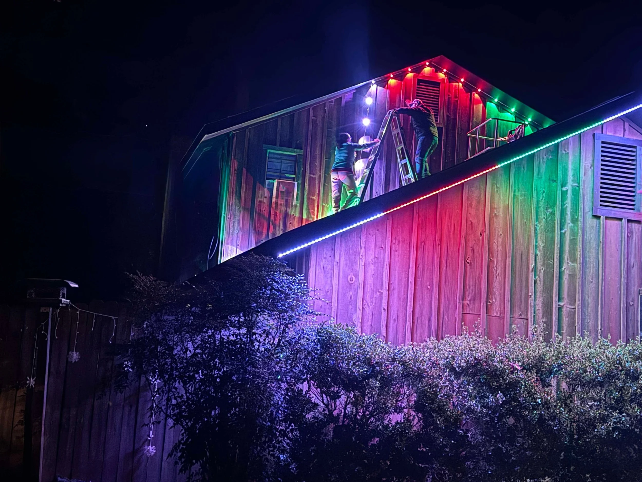 Two people decorating a wooden house with colorful Christmas lights at night, using a ladder.