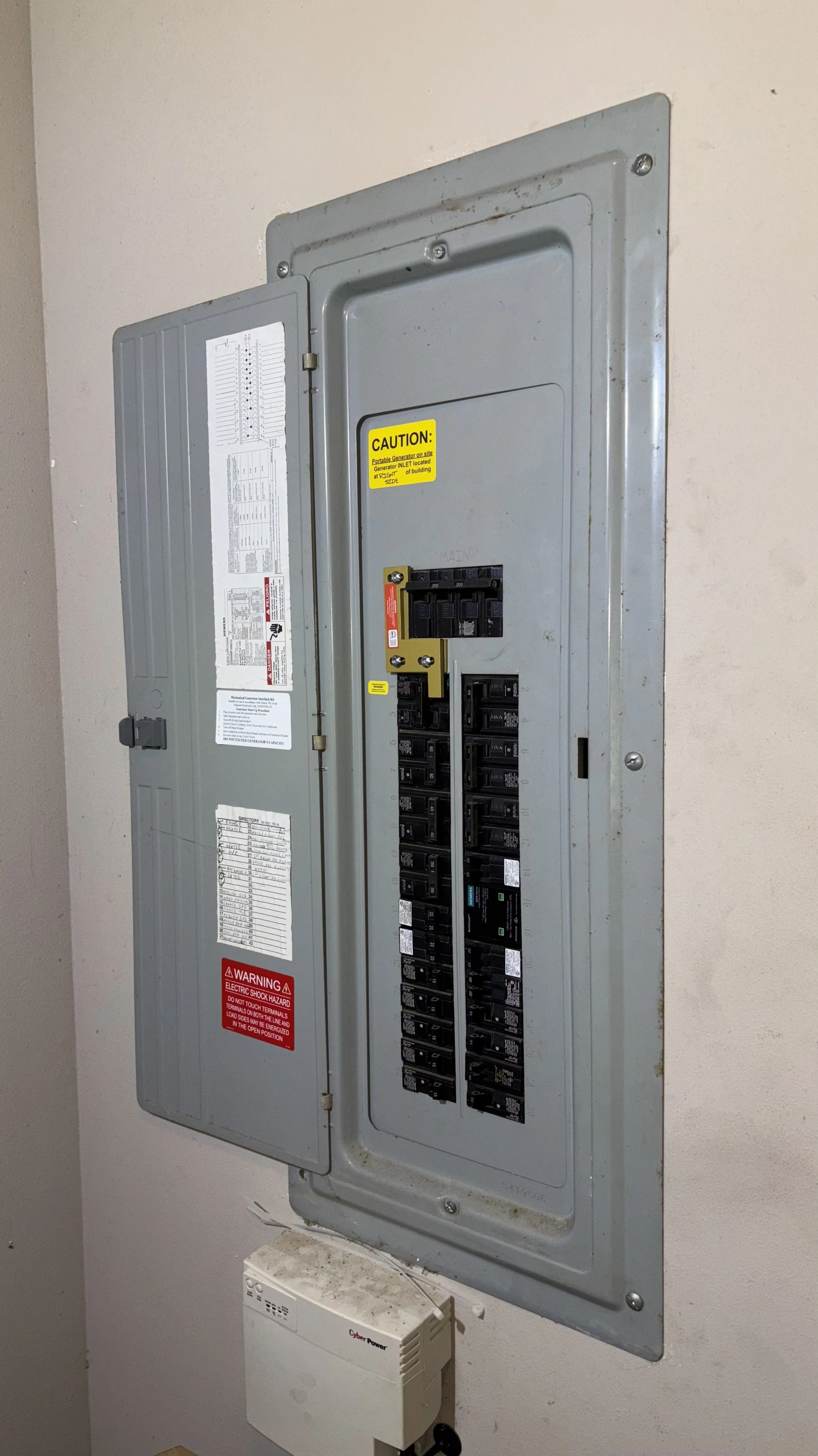 Electric breaker box with warning labels and a small white device mounted below it, attached to a beige wall.