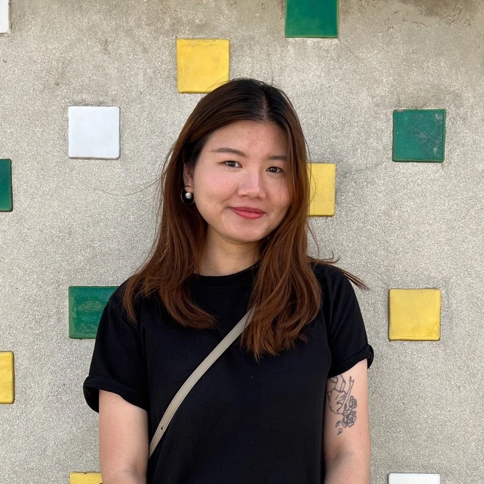 A young woman with medium-length brown hair, wearing a black t-shirt, pearl earrings, and a crossbody bag, standing in front of a textured wall with colorful square tiles in yellow, green, and white.