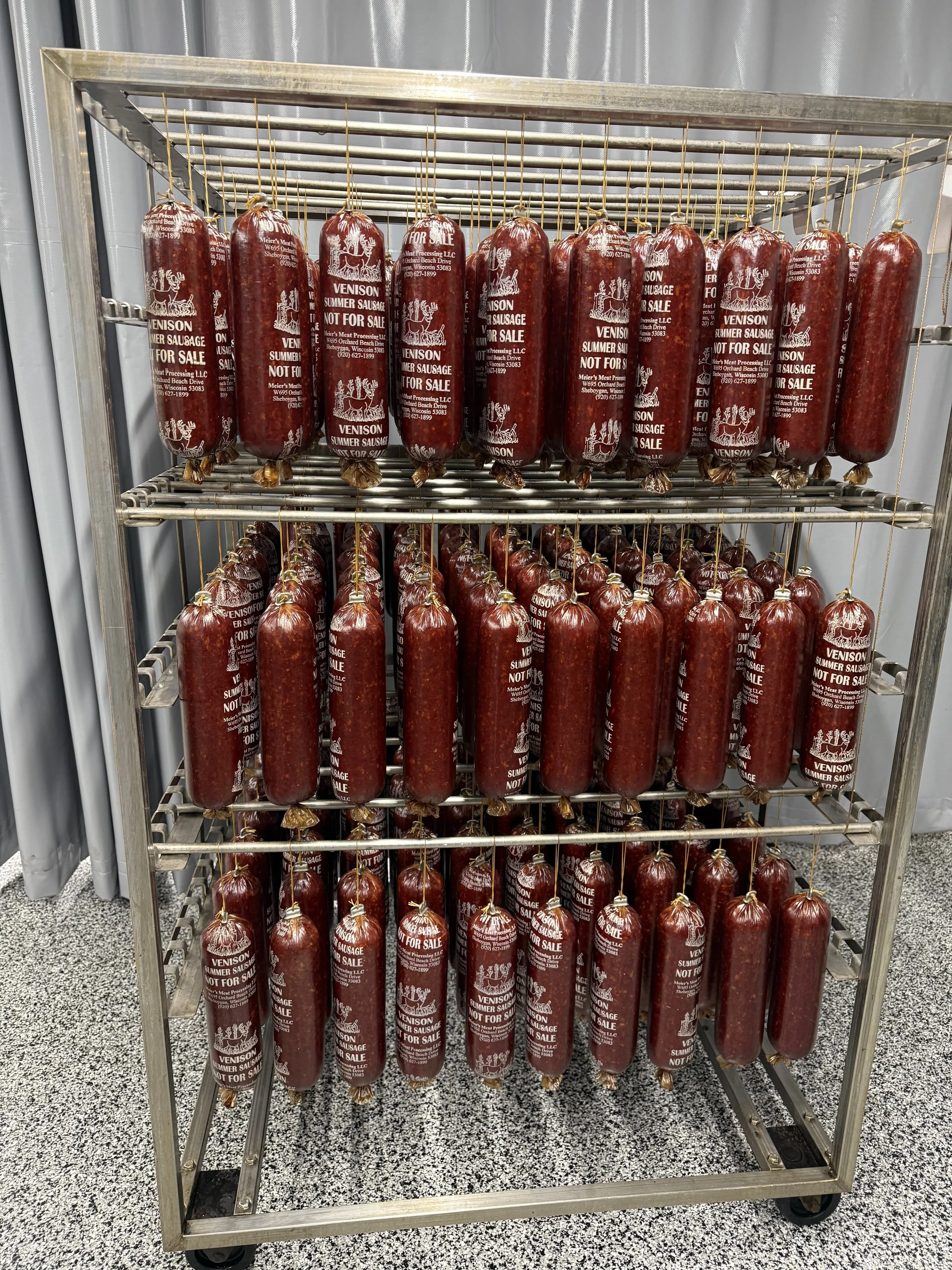 A metal rack filled with large packages of venison summer sausage on multiple shelves inside a room.