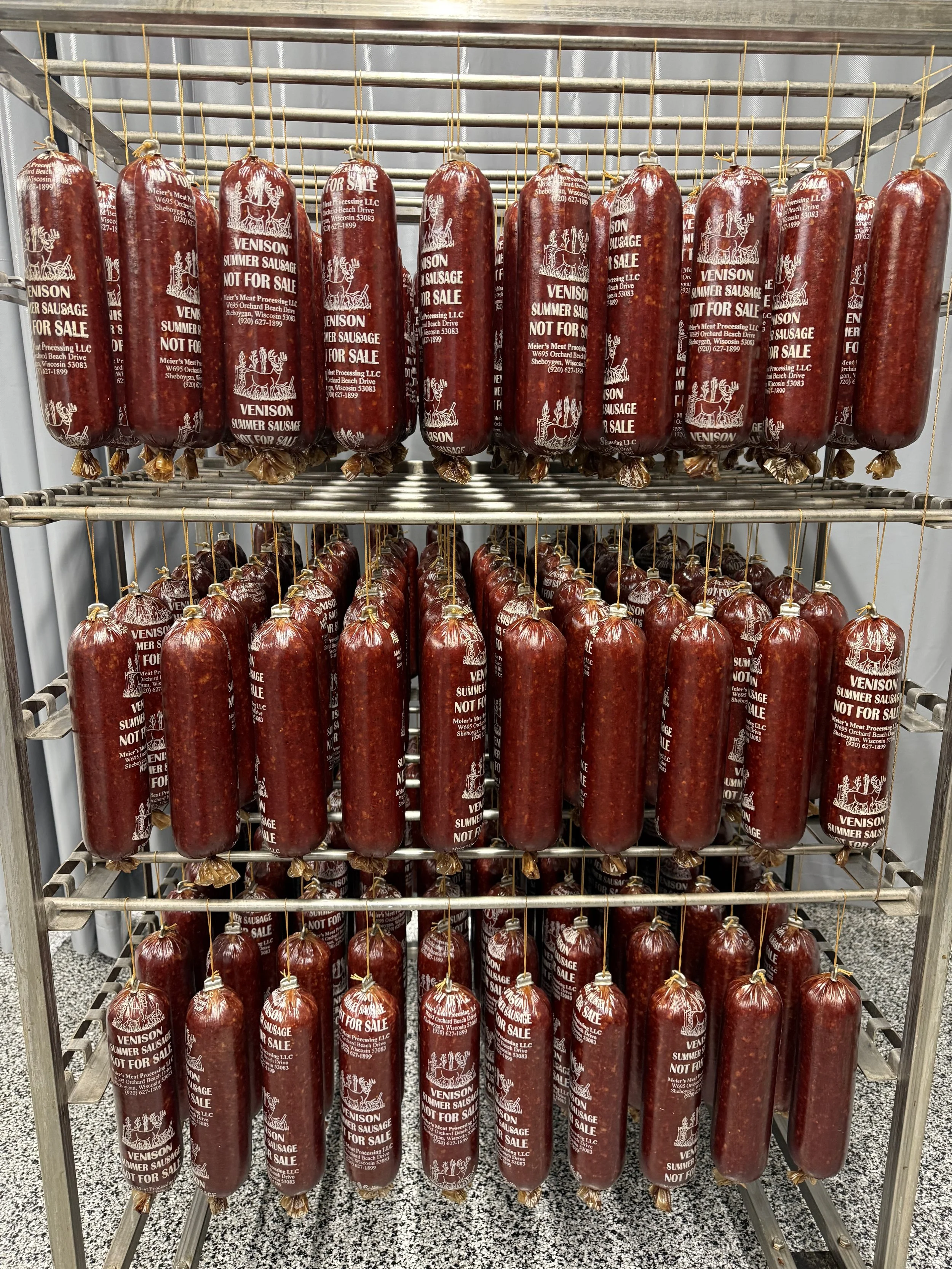 A metal rack holding multiple packages of venison summer sausage, all wrapped in red casing with white labels indicating they are not for sale, in a retail setting.