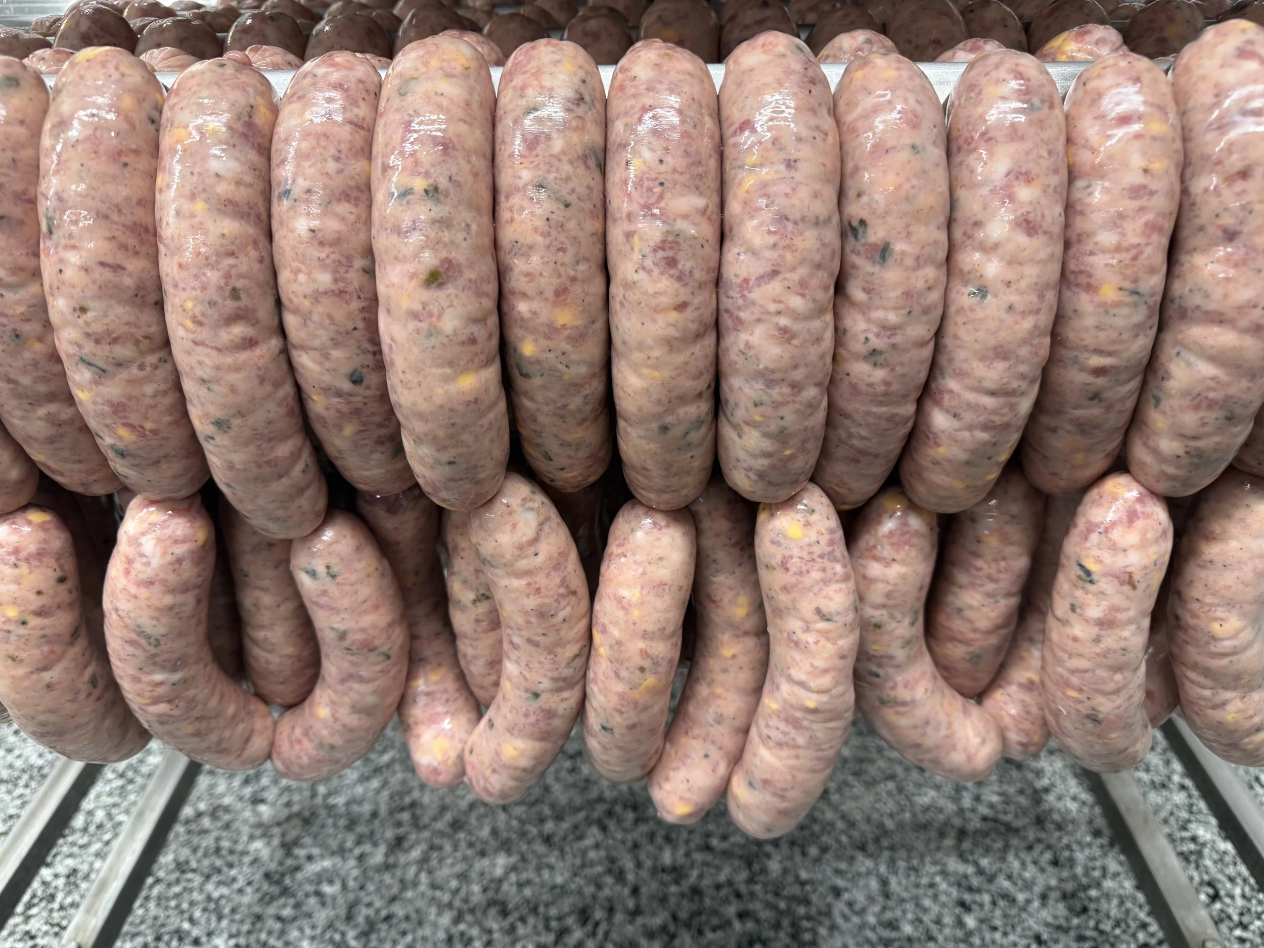 Multiple raw sausages with specks of herbs and spices hanging on a metal rack.