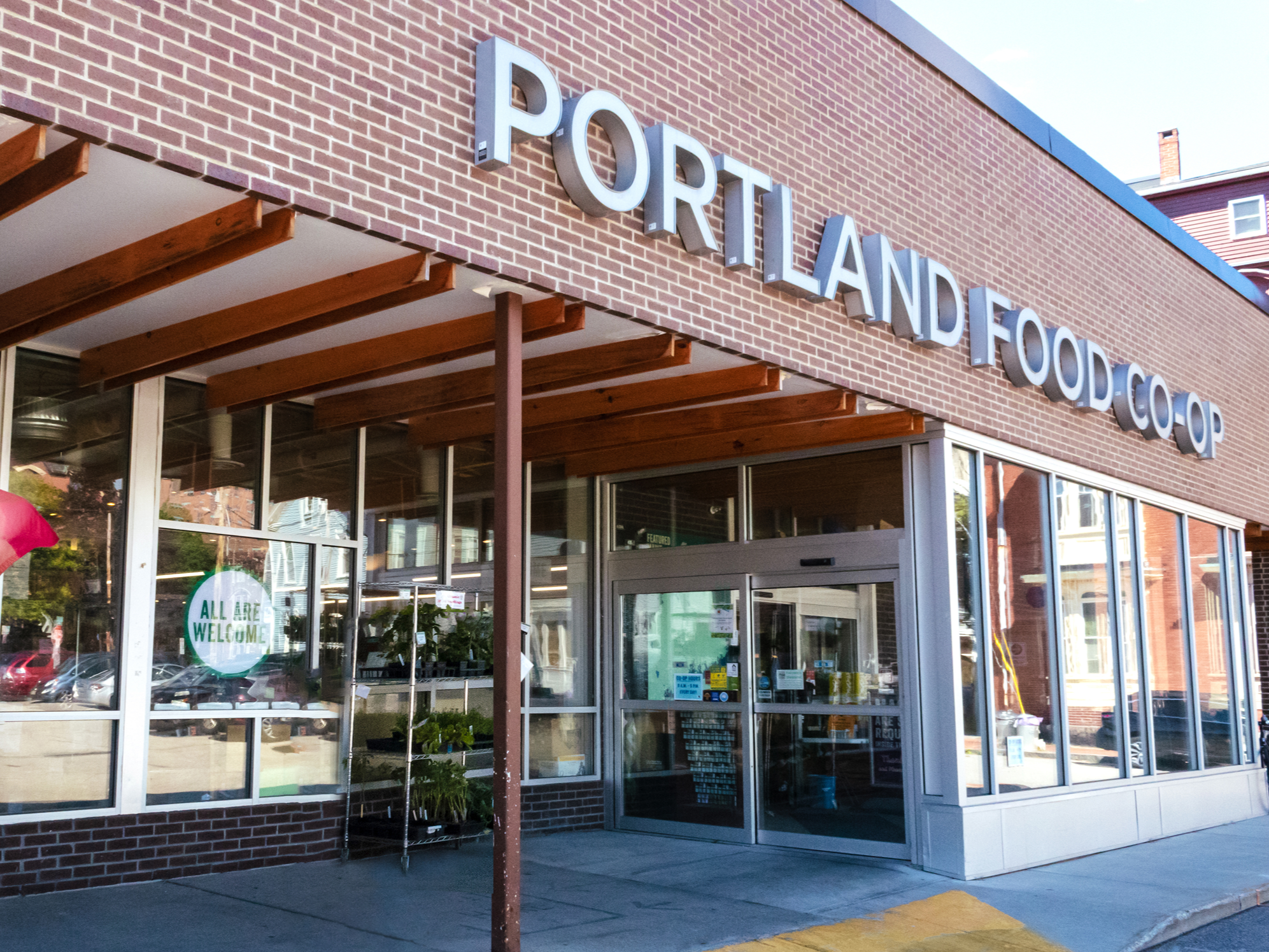Exterior of a grocery store called 'Portland Food Co-op' with large metal letters on the brick wall. Glass storefront showing reflection of parked cars and street scene. Wooden awning above the entrance. Sign in window reads 'All Are Welcome'.