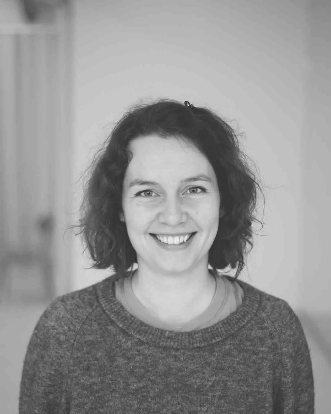 Zwart-wit foto van een lachende vrouw met donker, golvend haar, gekleed in een grijze trui, staand in een lichte kamer.