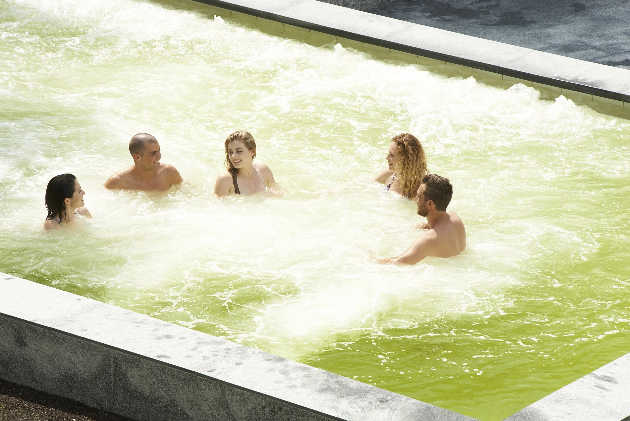 Sechs Menschen befinden sich in einem Pool mit grünlichem Wasser, sie unterhalten sich und haben Spaß.