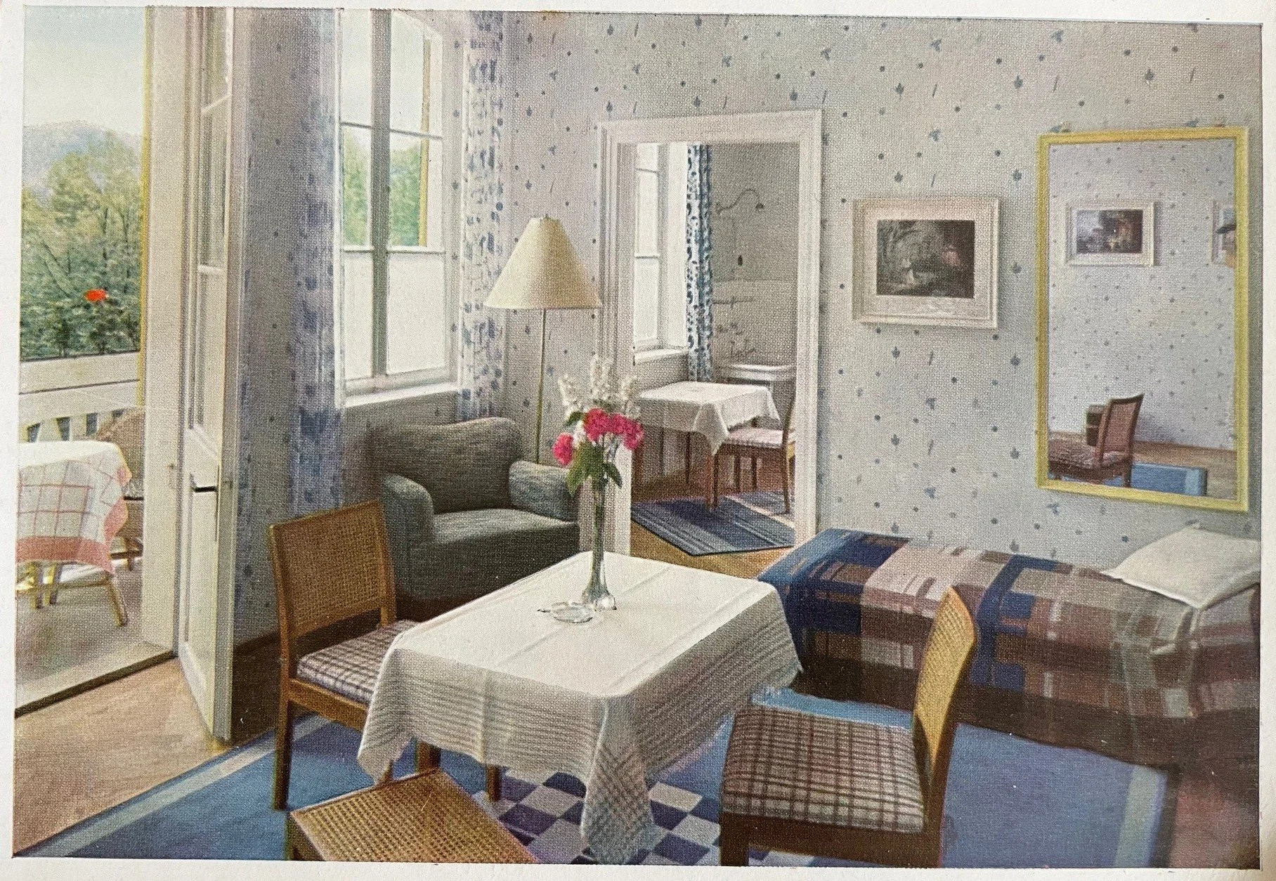 Ein Hotelzimmer im Stil der 50er Jahre, mit Polkatapete, ein Tisch mit weißen Tischtuch und Blumen, Sofas, Stühlen und Wandbildern, Blick auf einen Balkon im Freien.