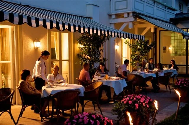 Abendliche Szene eines Restaurants im Freien mit mehreren Tischen, an denen Gäste speisen und ein Kellner serviert. Es ist warmes Licht, und im Vordergrund sind Blumen und Kerzen zu sehen.